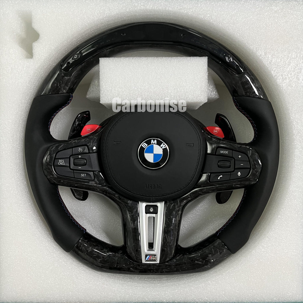 Custom BMW M performance steering wheel featuring a forged carbon fiber finish, black smooth leather side grips, and vibrant red M1 and M2 button accents.