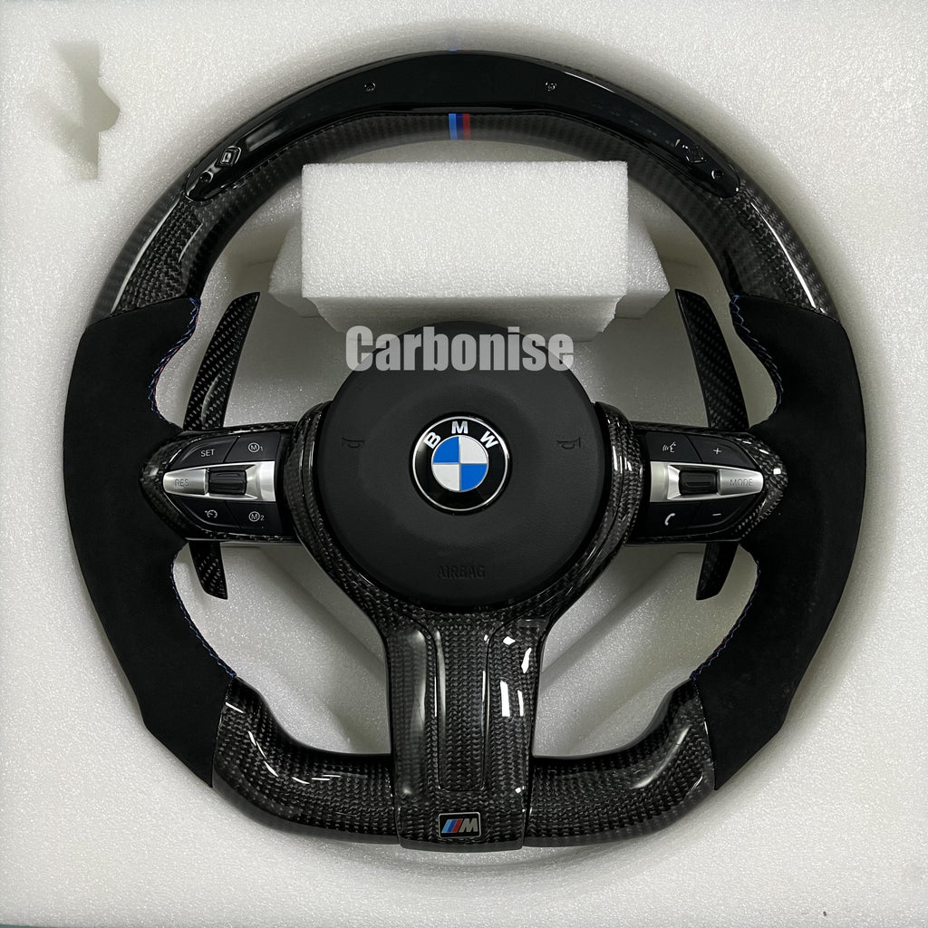 Custom BMW M performance steering wheel featuring high-gloss carbon fiber, black Alcantara grips, blue contrast stitching, and a blue top racing stripe.