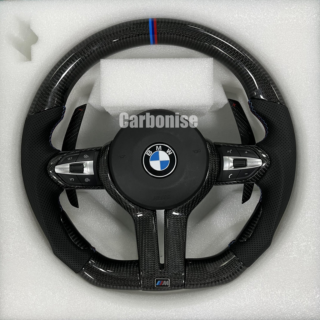 Custom BMW M Performance steering wheel featuring high-gloss twill carbon fiber, a signature blue, light blue, and red M-tri color top racing stripe, and black perforated leather side grips with blue contrast stitching.
