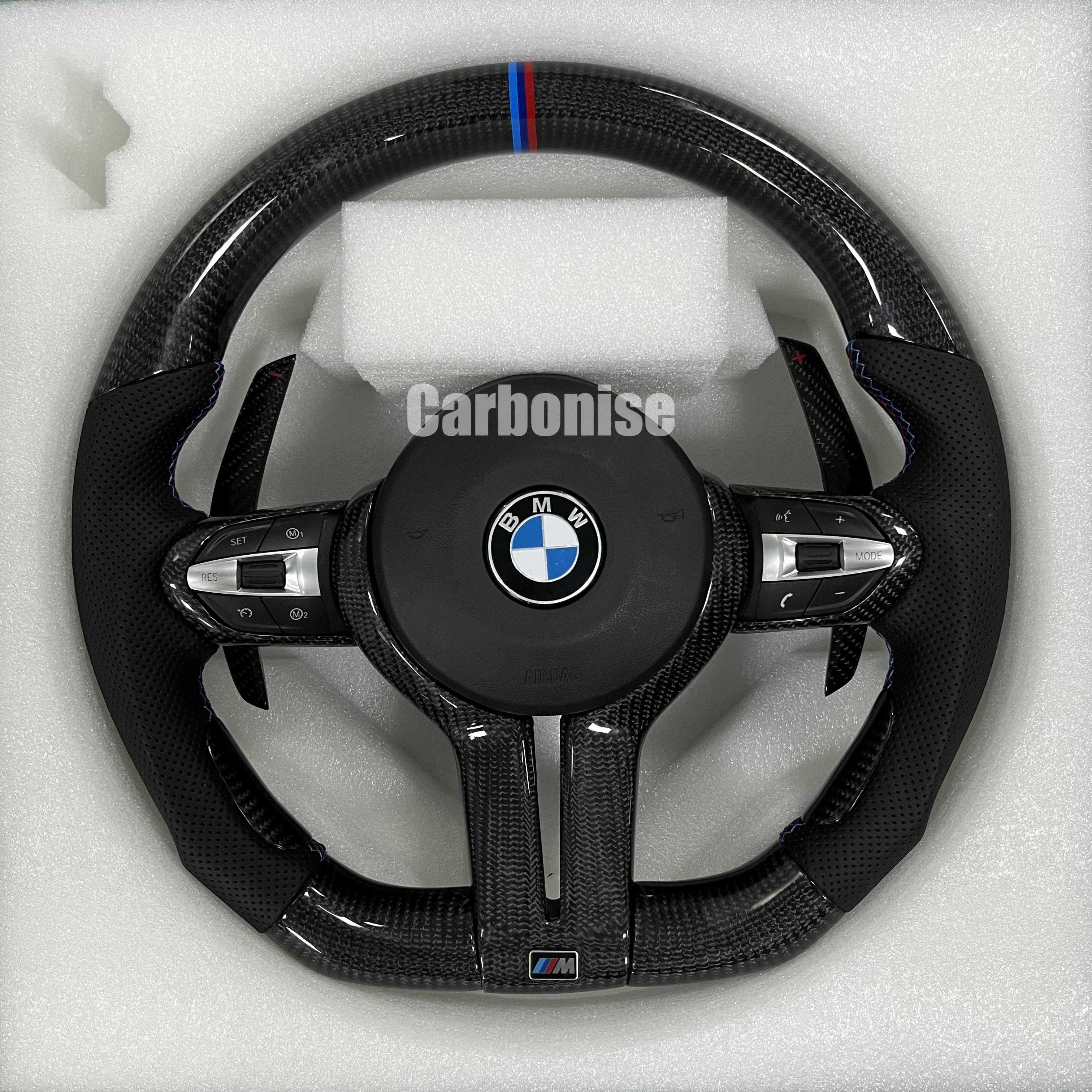 Custom BMW M Performance steering wheel featuring high-gloss twill carbon fiber, a signature blue, light blue, and red M-tri color top racing stripe, and black perforated leather side grips with blue contrast stitching.