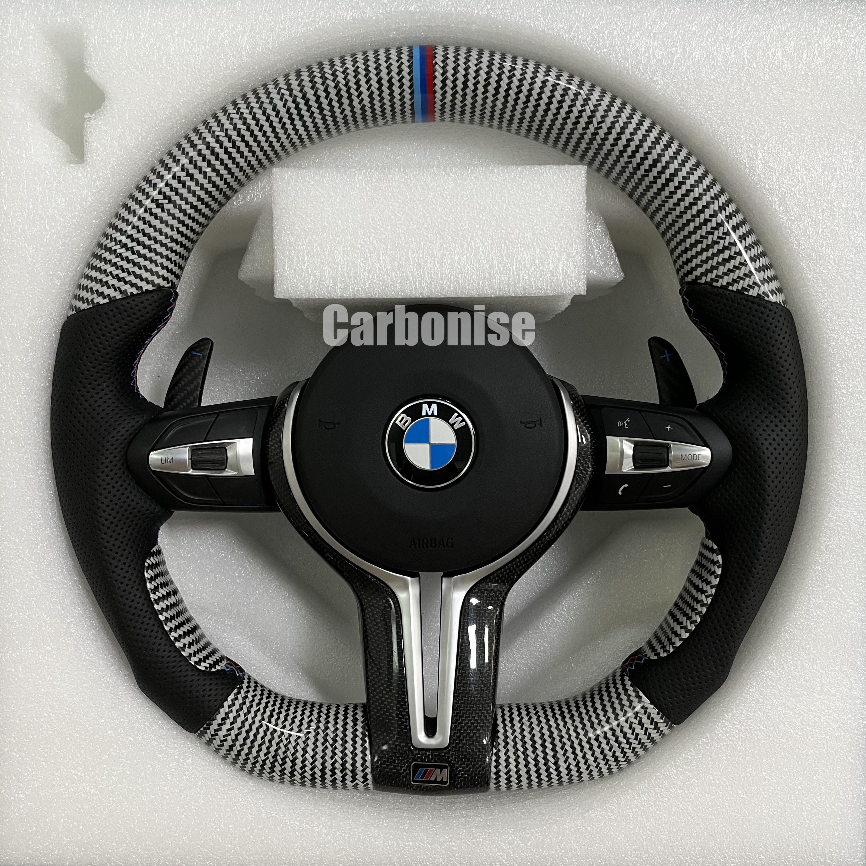 Custom BMW M Performance steering wheel featuring silver twill carbon fiber, a signature M-tri color top racing stripe, and black perforated leather side grips with blue contrast stitching.