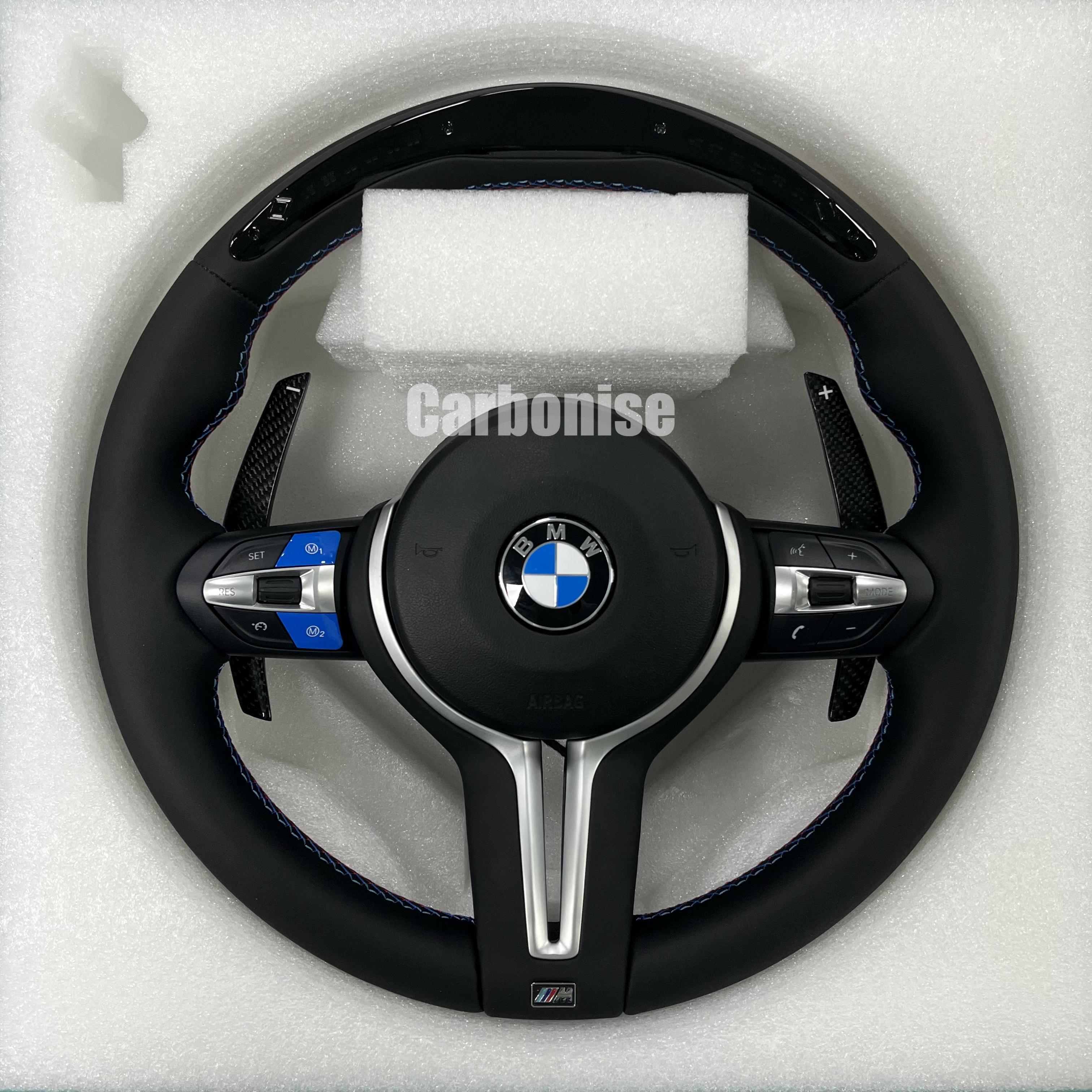Custom BMW M Performance steering wheel featuring a gloss carbon fiber top, black Alcantara side grips with blue stitching, and blue-anodized M1/M2 button accents on the steering wheel controls.