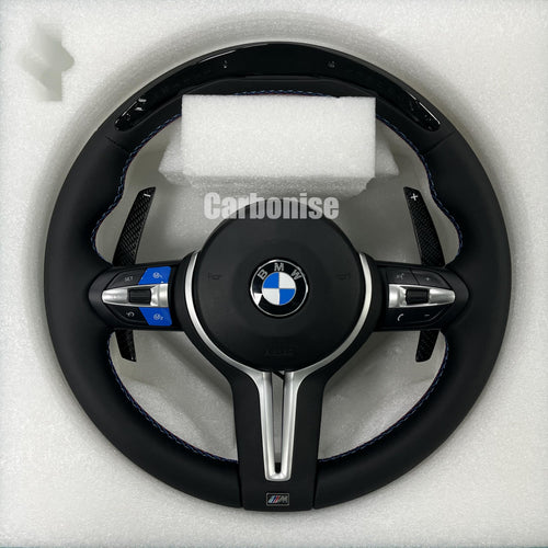 Custom BMW M Performance steering wheel featuring a gloss carbon fiber top, black Alcantara side grips with blue stitching, and blue-anodized M1/M2 button accents on the steering wheel controls.
