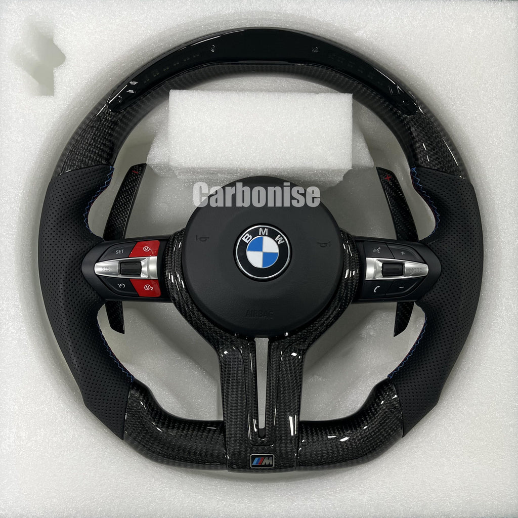 Custom BMW M performance steering wheel featuring gloss carbon fiber, black perforated leather side grips with blue stitching, and red button accents.