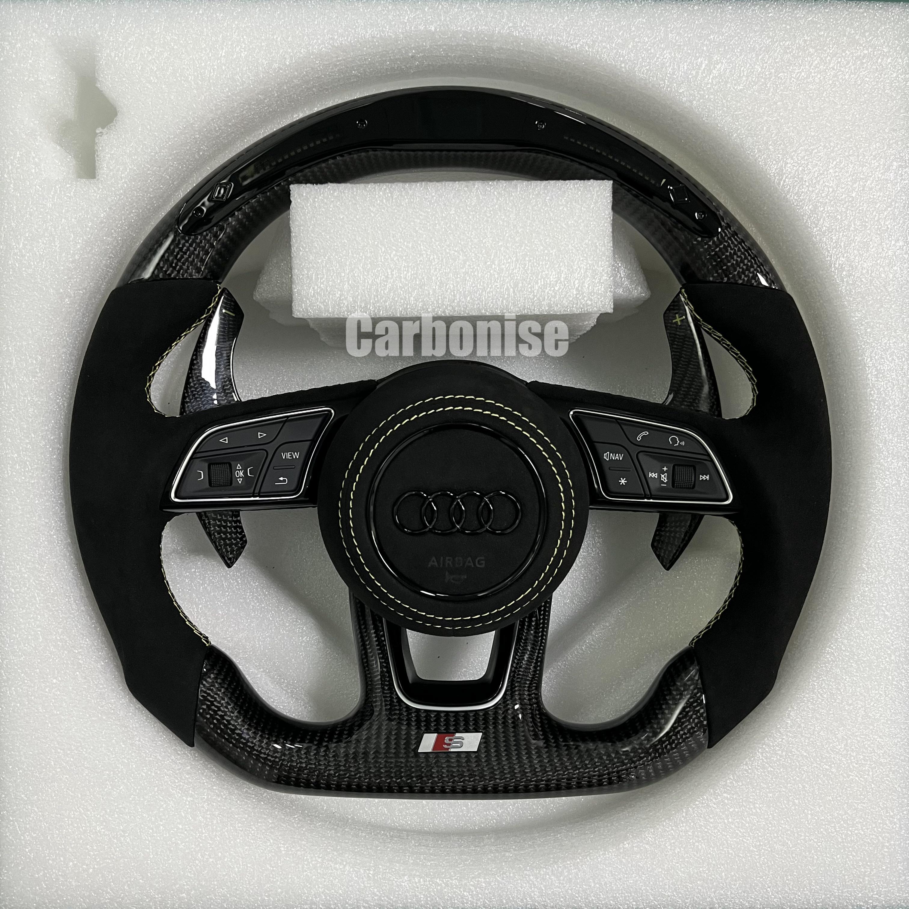 Custom Audi S-Line steering wheel featuring gloss carbon fiber, black Alcantara side grips with yellow stitching, and a blacked-out Audi rings emblem on a leather airbag cover.