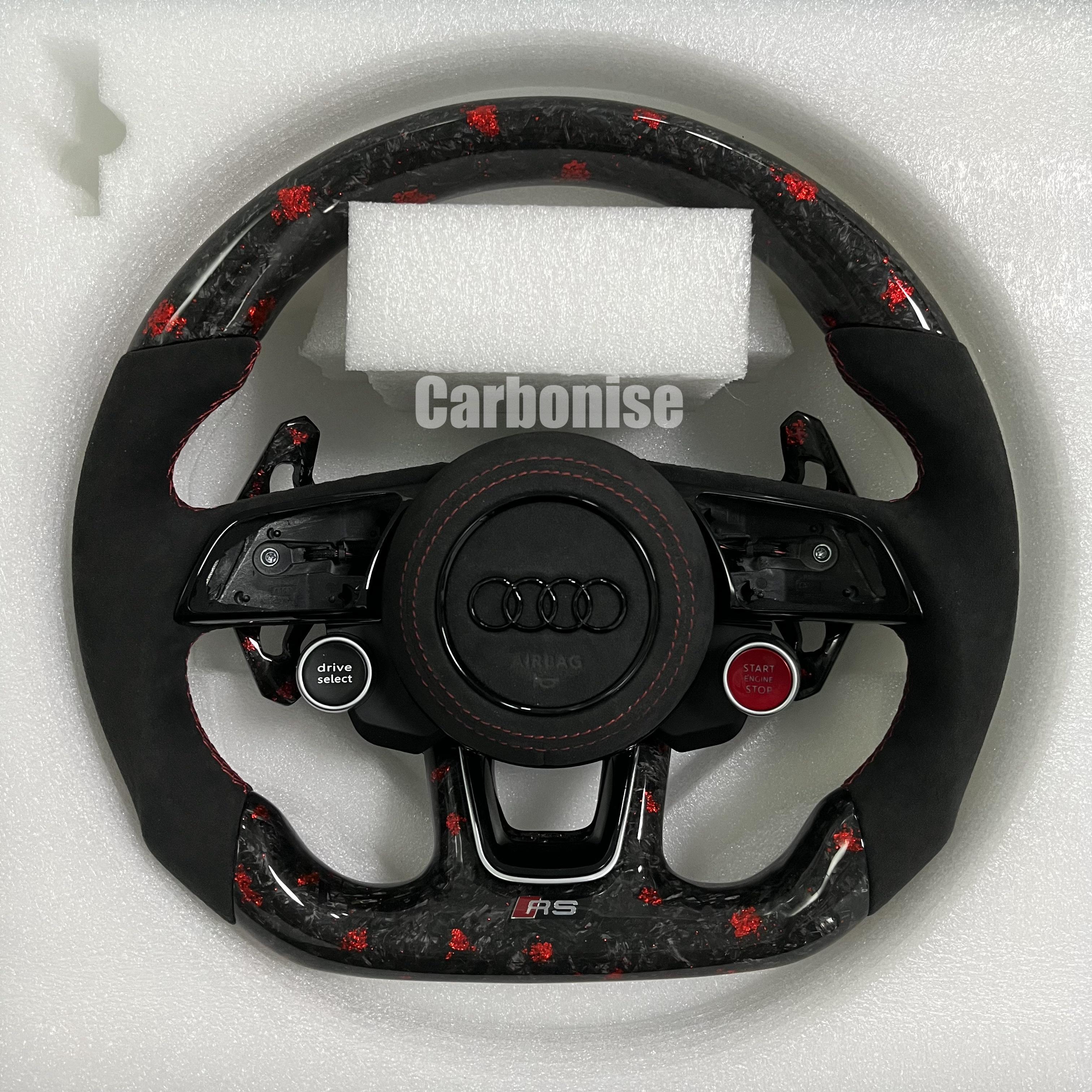 Custom Audi RS steering wheel in forged carbon fiber with red flakes, featuring black Alcantara side grips, red stitching, and integrated red Start/Stop and black Drive Select buttons.