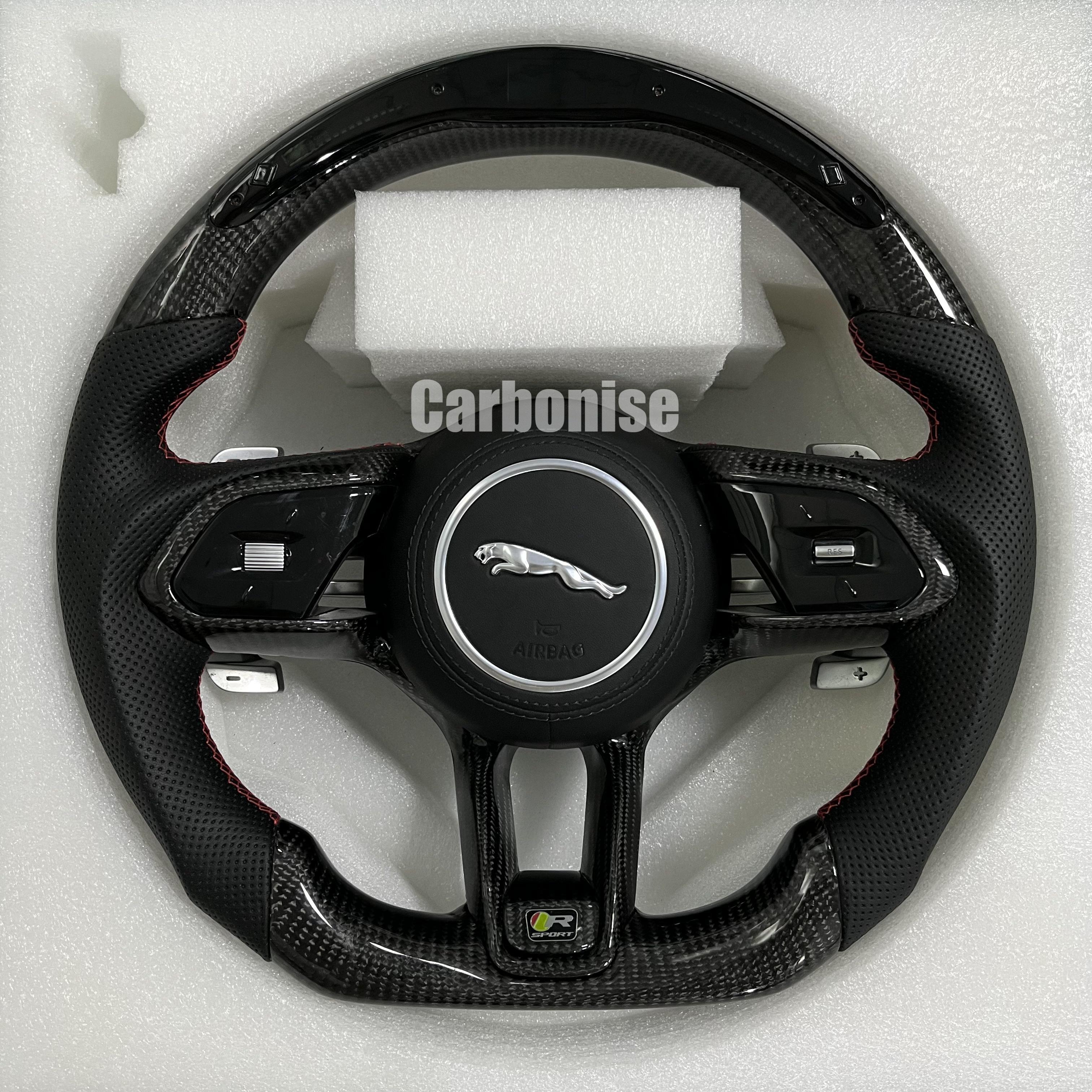 Custom Jaguar carbon fibre steering wheel with perforated black leather grips, red stitching, integrated paddle shifters, and flat-bottom performance design.