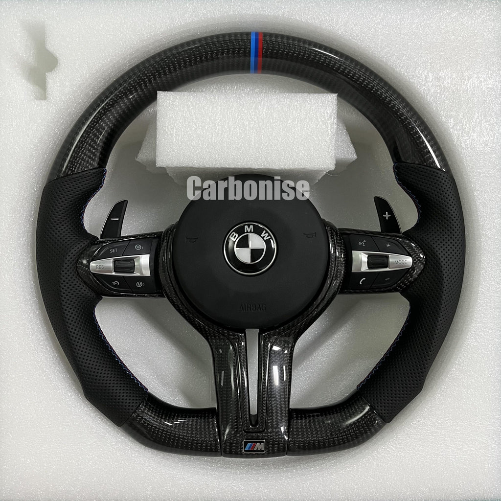 Custom BMW M Performance steering wheel featuring high-gloss twill carbon fiber, a signature blue, light blue, and red M-tri color top racing stripe, and black perforated leather side grips with blue contrast stitching.