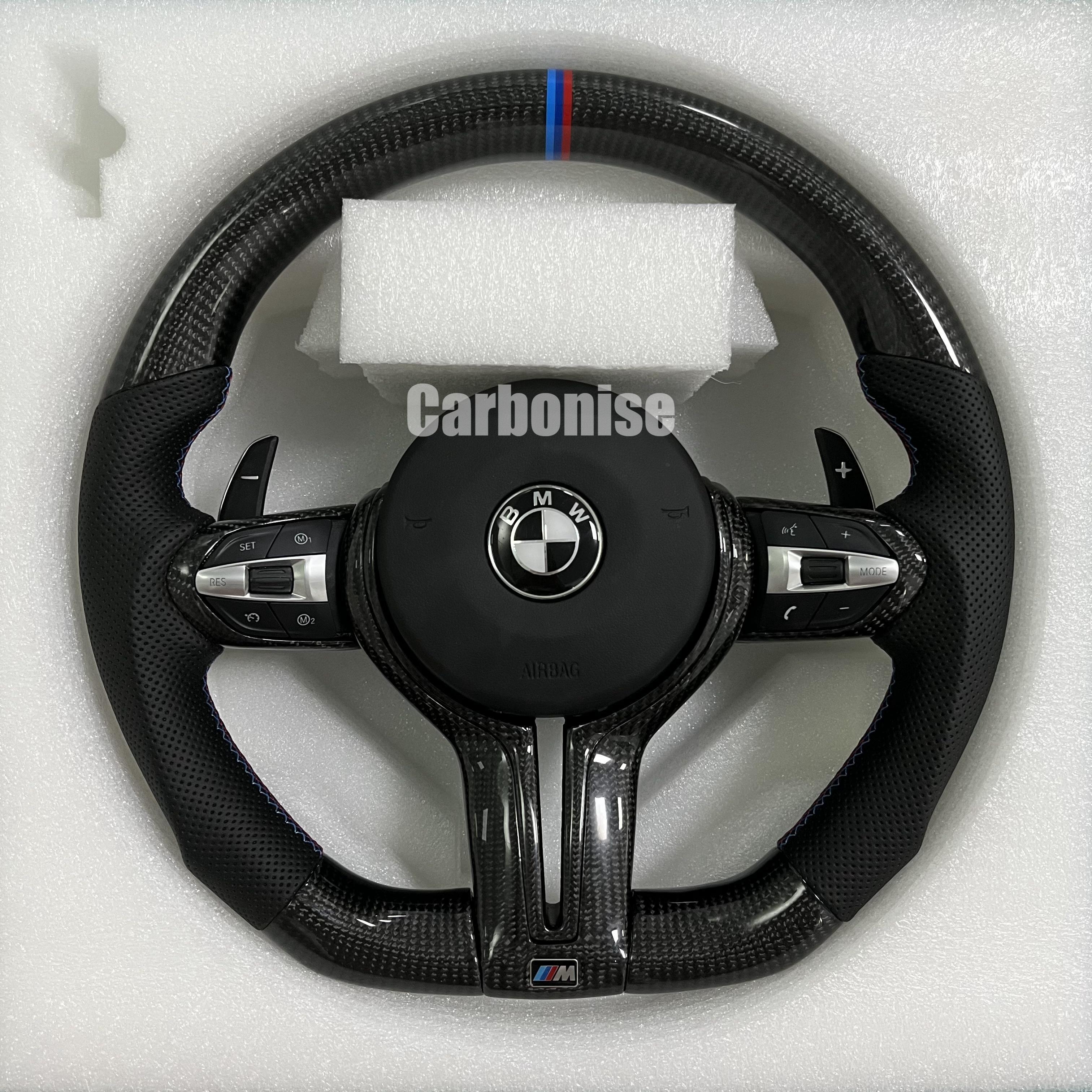 Custom BMW M Performance steering wheel featuring high-gloss twill carbon fiber, a signature blue, light blue, and red M-tri color top racing stripe, and black perforated leather side grips with blue contrast stitching.