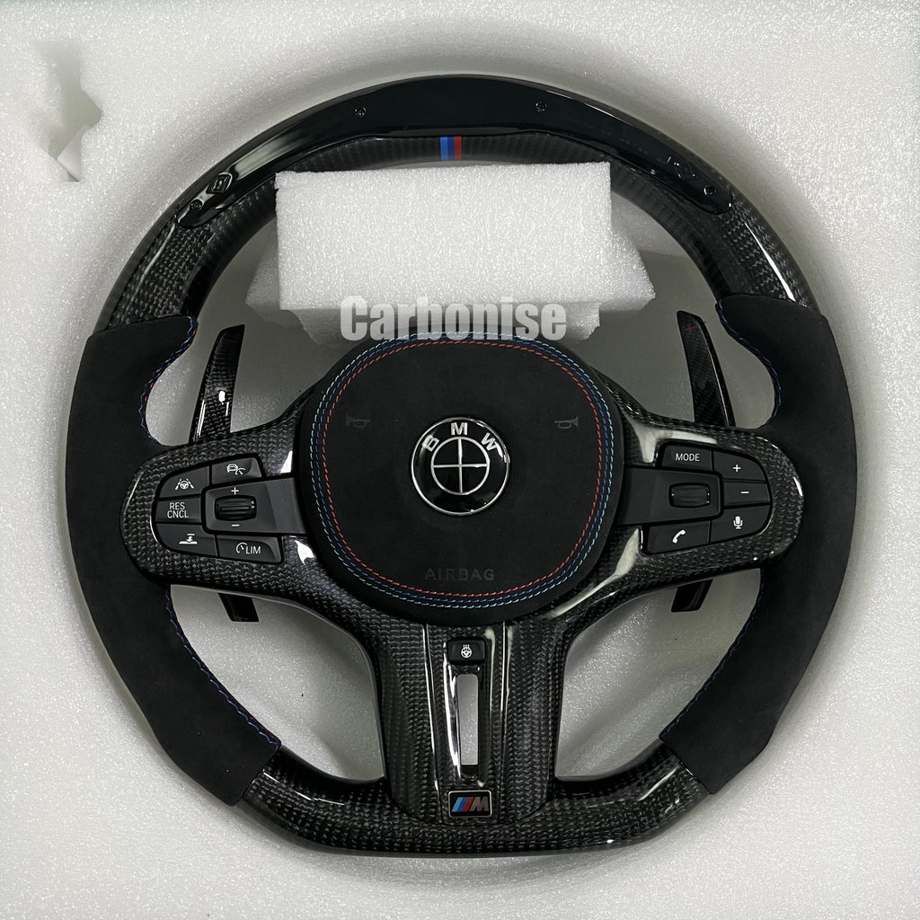Custom BMW M performance steering wheel featuring high-gloss carbon fiber, black Alcantara side grips with blue stitching, and a signature M-tri color top racing stripe.