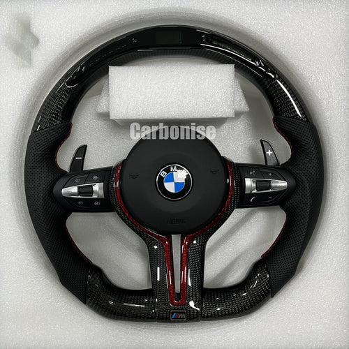 Custom BMW M steering wheel featuring gloss carbon fiber with a digital LED shift display, black perforated leather grips, and red contrast accents.