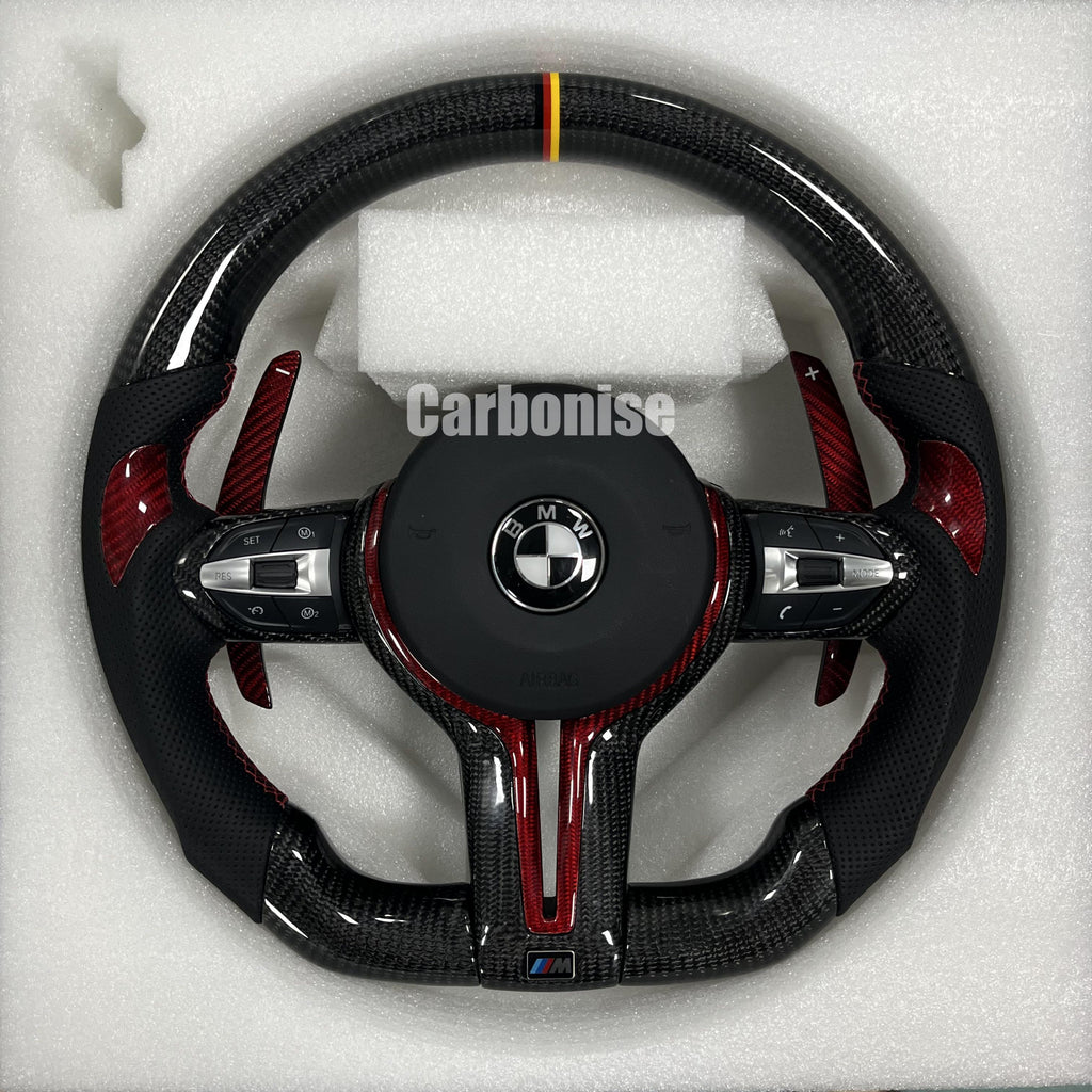 Custom BMW M steering wheel with gloss carbon fiber, a German flag racing stripe, red tinted carbon accents, and perforated leather grips with red stitching.