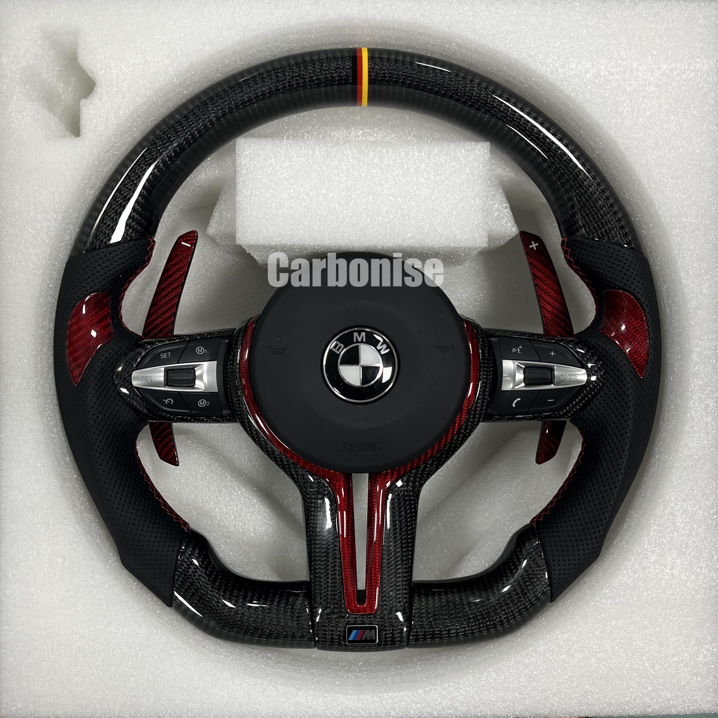 Custom BMW M steering wheel with gloss carbon fiber, a German flag racing stripe, red tinted carbon accents, and perforated leather grips with red stitching.