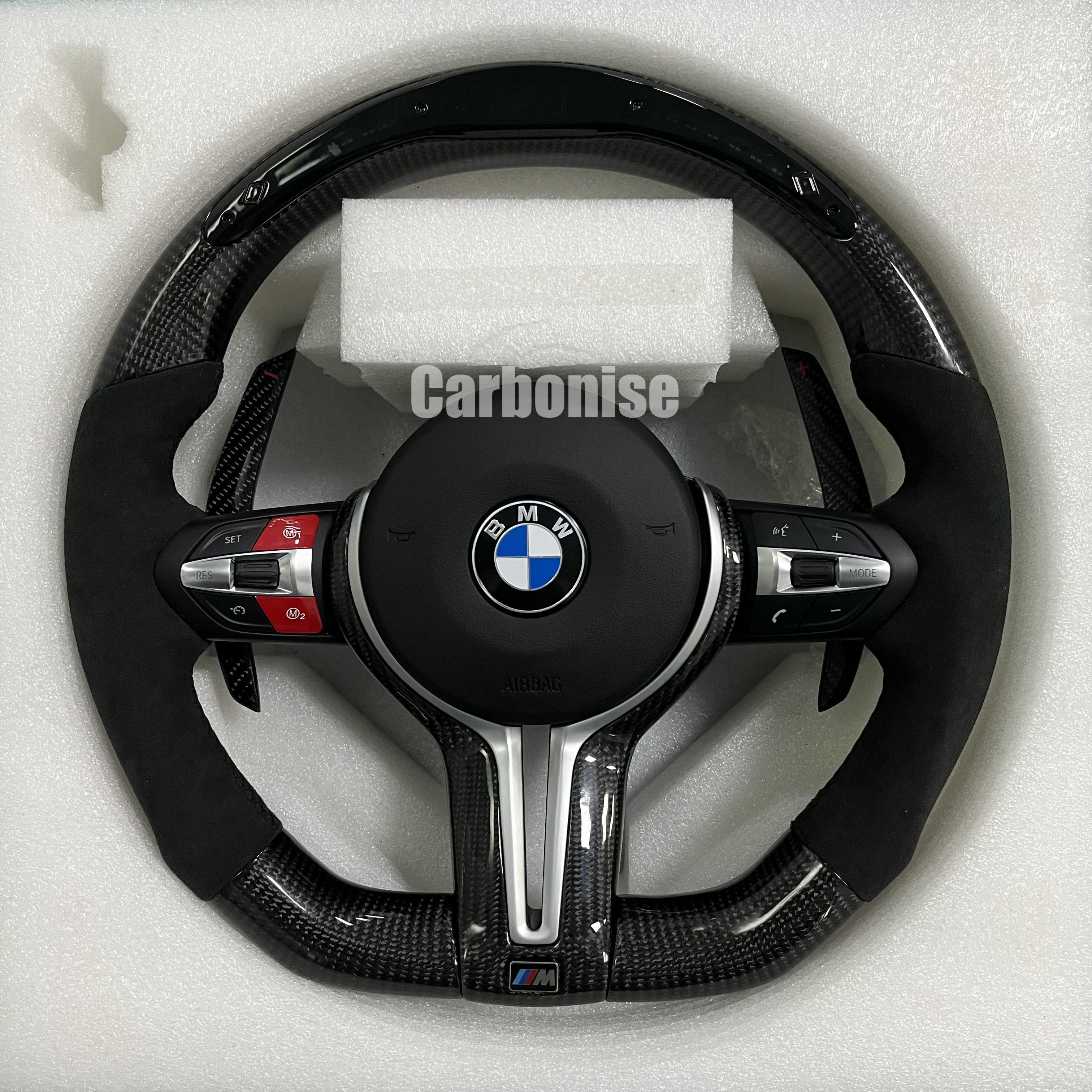 Custom BMW M performance steering wheel featuring gloss carbon fiber, black perforated leather side grips with blue stitching, and red button accents.
