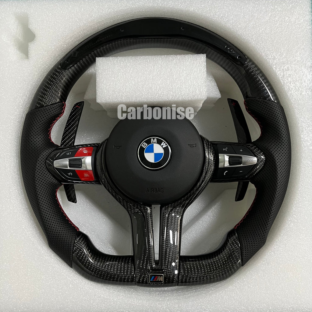 Custom BMW M performance steering wheel featuring gloss carbon fiber, black perforated leather side grips with red stitching, and red button accents.