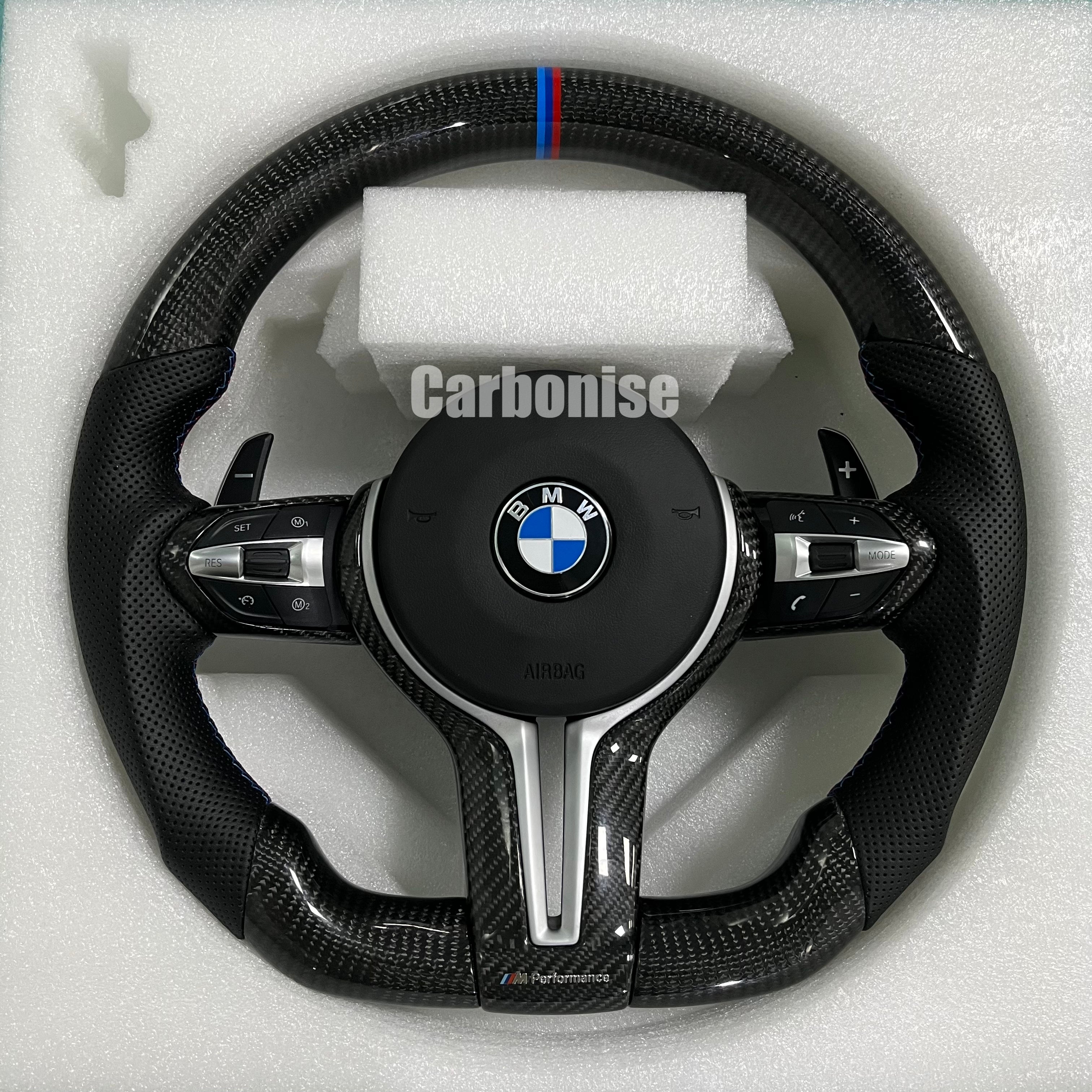 BMW M Performance steering wheel featuring gloss carbon fiber, a signature blue, light blue, and red M-tri color top racing stripe, and black perforated leather side grips with blue stitching.