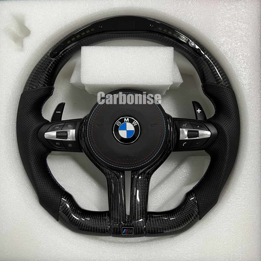 Custom BMW M performance steering wheel featuring gloss carbon fiber with a digital LED shift display, black perforated leather grips, and blue stitching.
