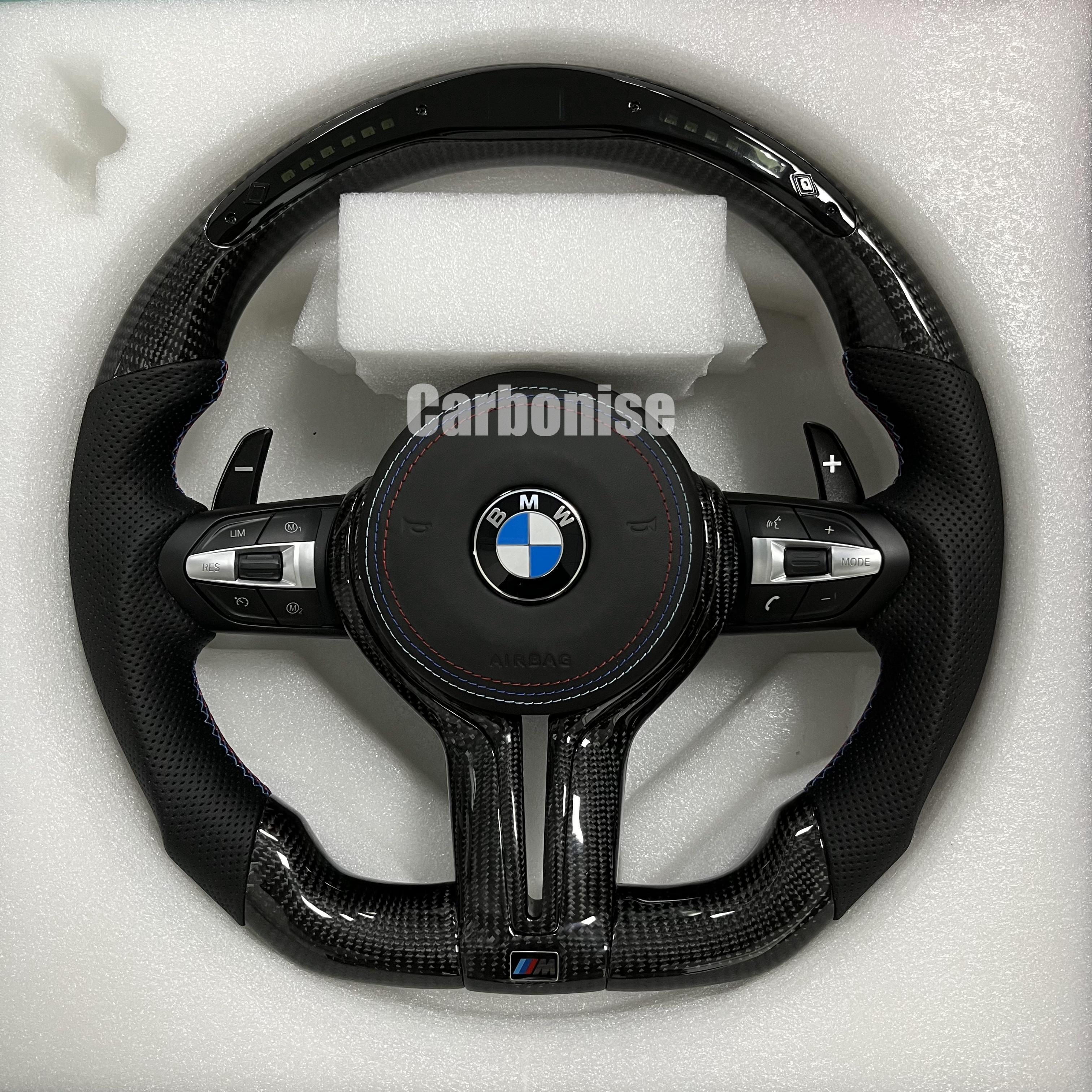 Custom BMW M performance steering wheel featuring gloss carbon fiber with a digital LED shift display, black perforated leather grips, and blue stitching.