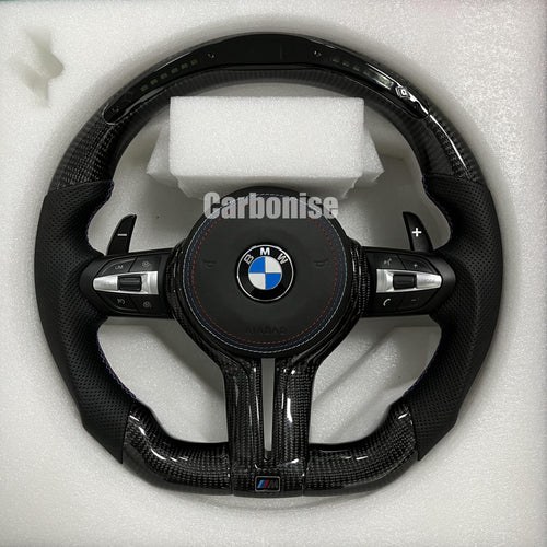 Custom BMW M performance steering wheel featuring gloss carbon fiber with a digital LED shift display, black perforated leather grips, and blue stitching.