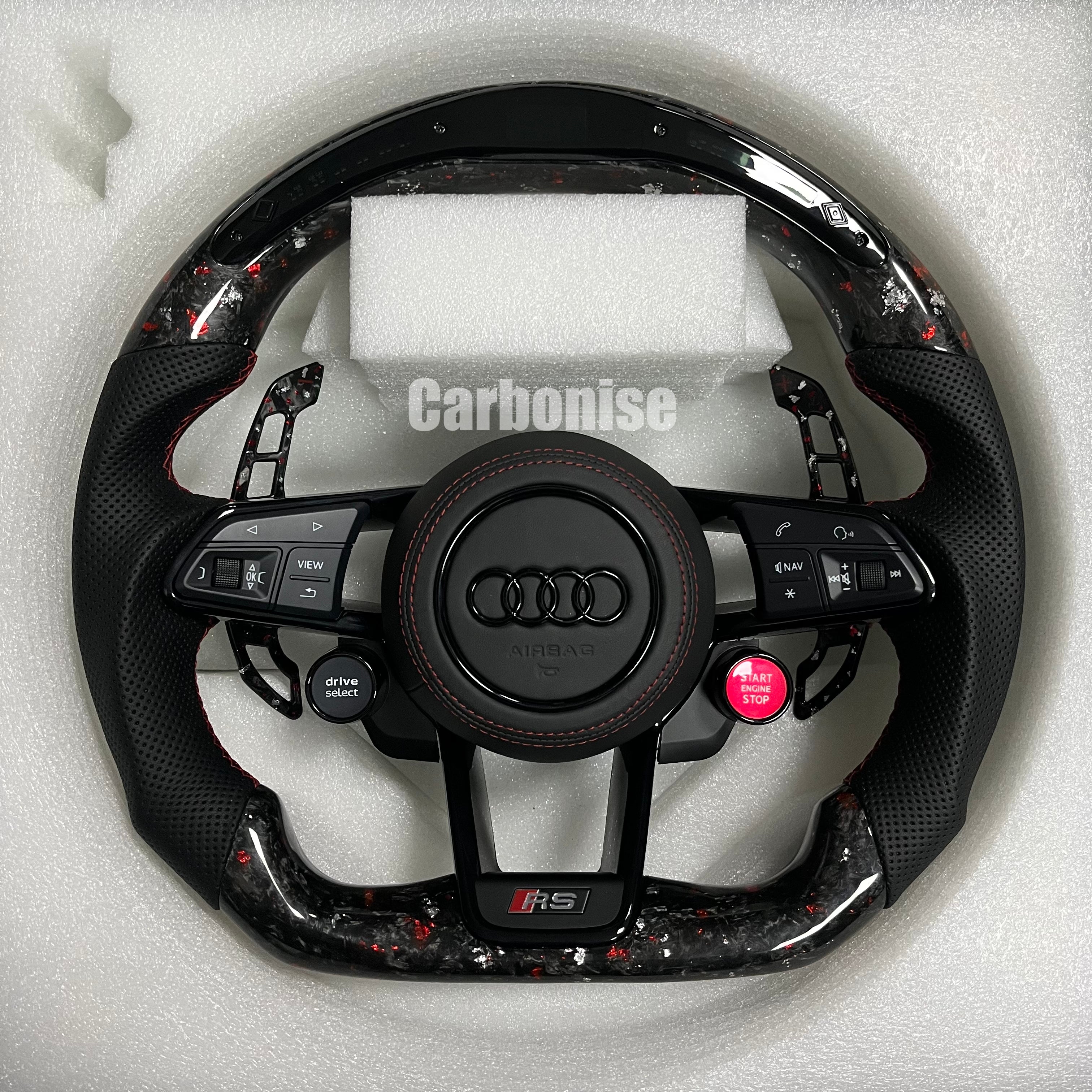 Custom Audi RS steering wheel in forged carbon fiber with red flakes, featuring black perforated leather grips, red stitching, and integrated red Start/Stop and black Drive Select buttons.