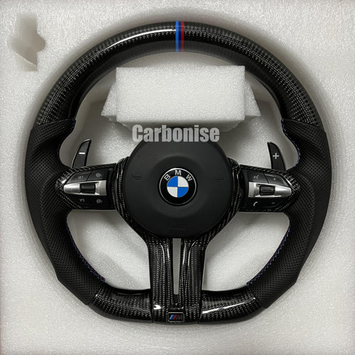 Custom BMW M Performance steering wheel featuring gloss carbon fiber, a signature blue, light blue, and red M-tri color top racing stripe, and black perforated leather side grips with blue stitching.