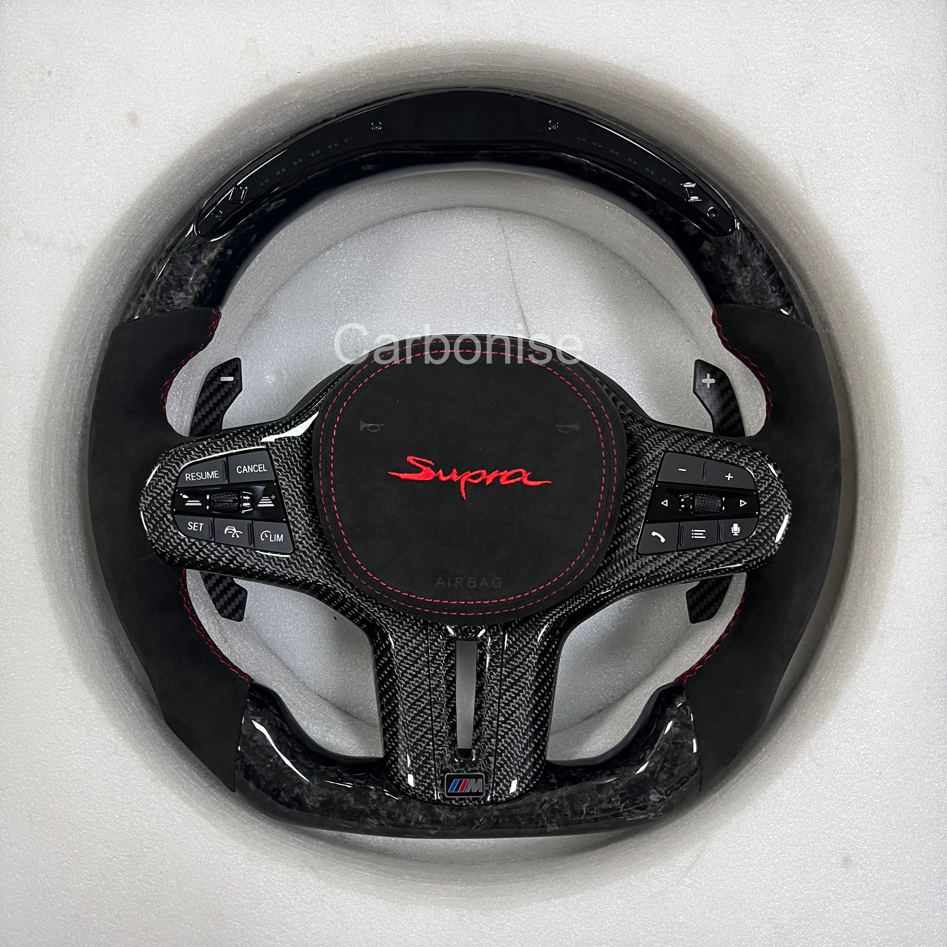  Supra steering wheel with carbon fibre top and bottom, Alcantara side grips, red Supra embroidery, red stitching, and carbon paddle shifters. Led shift light