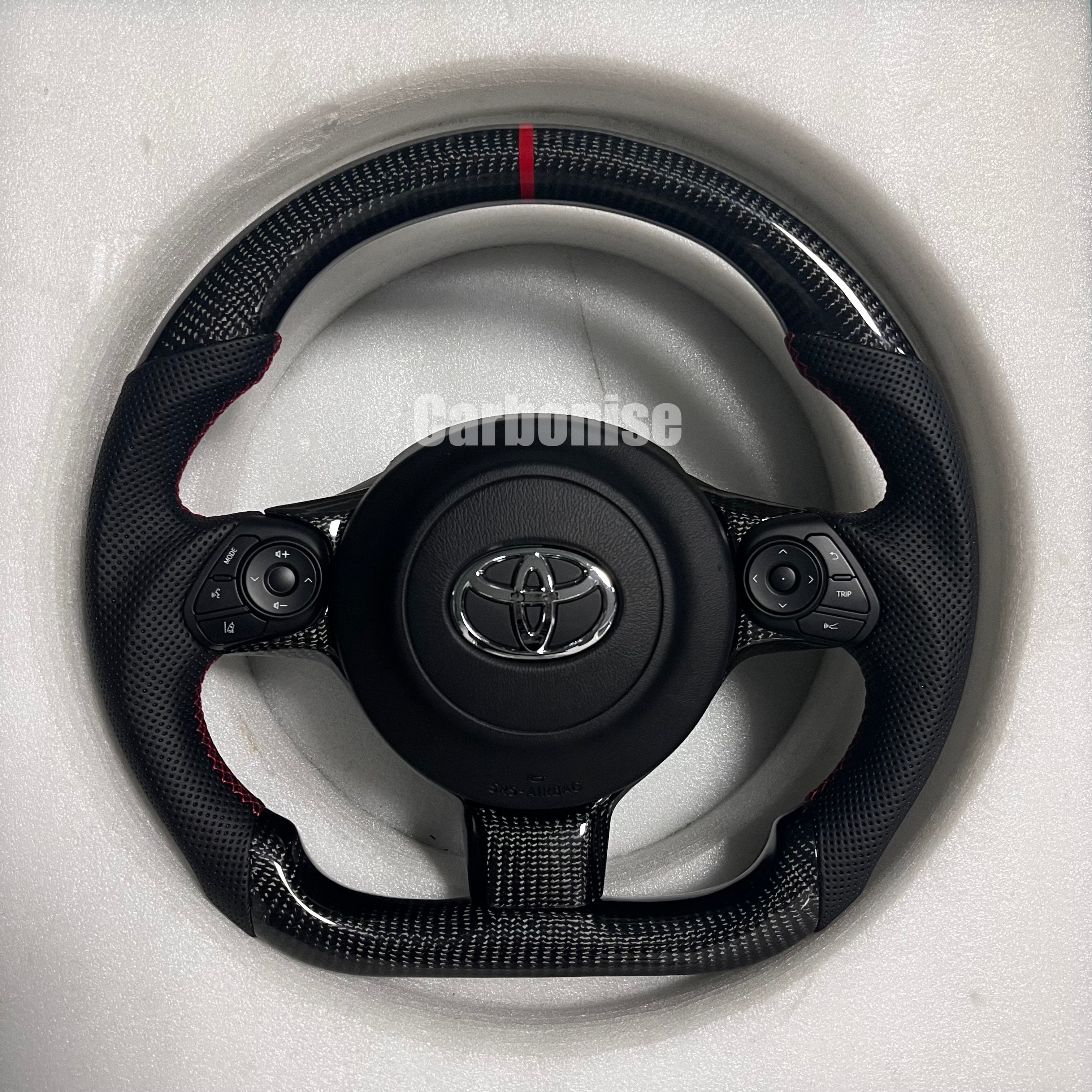 Carbonise carbon fiber Toyota steering wheel with red center stripe, perforated leather grips, OEM buttons, and flat bottom sport design