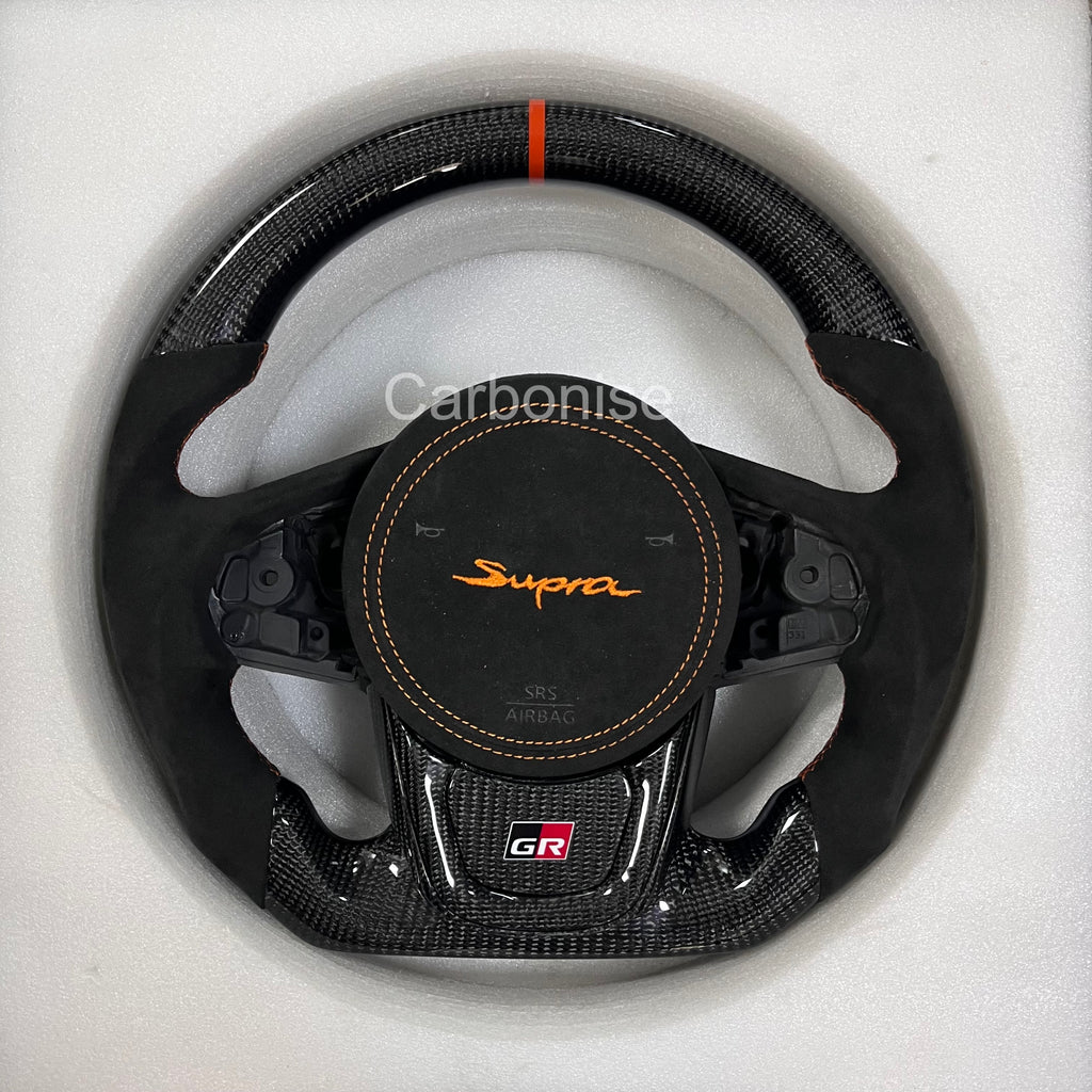Carbonise carbon fiber Toyota Supra GR steering wheel with orange center stripe, Alcantara grips, custom Supra airbag embroidery, and flat bottom design