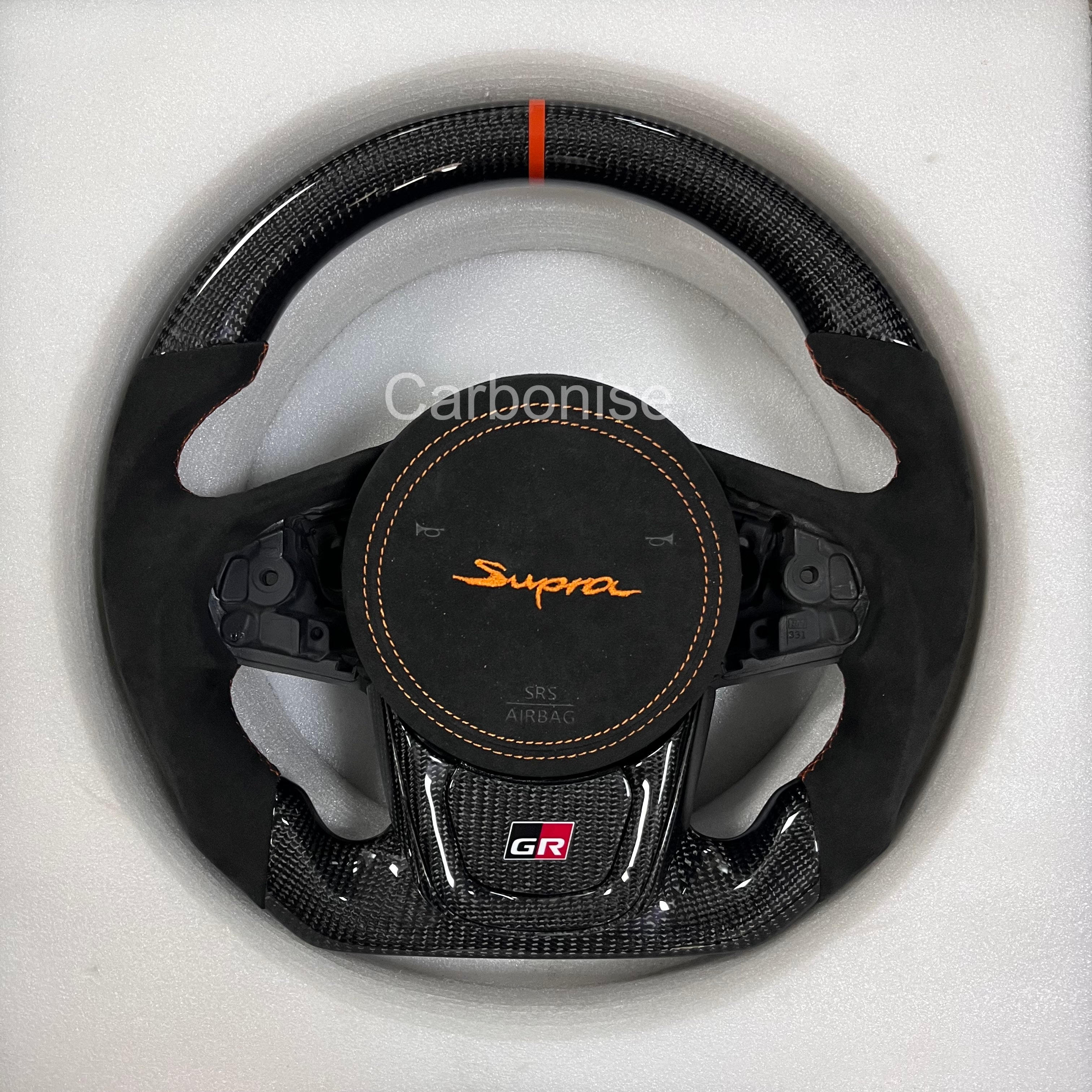 Carbonise carbon fiber Toyota Supra GR steering wheel with orange center stripe, Alcantara grips, custom Supra airbag embroidery, and flat bottom design