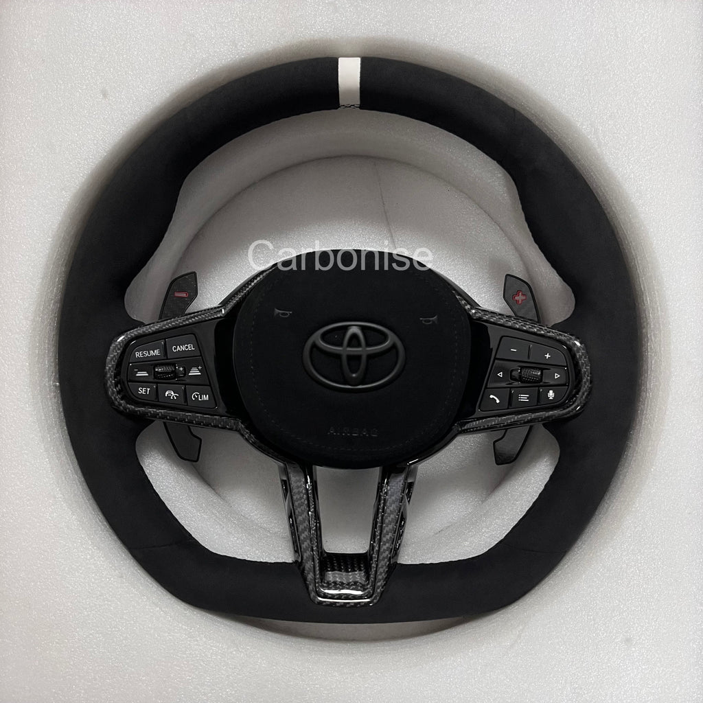 Carbonise Supra GR Alcantara steering wheel with white center stripe, carbon fiber trim, flat bottom design, paddle shifters, and OEM multifunction buttons