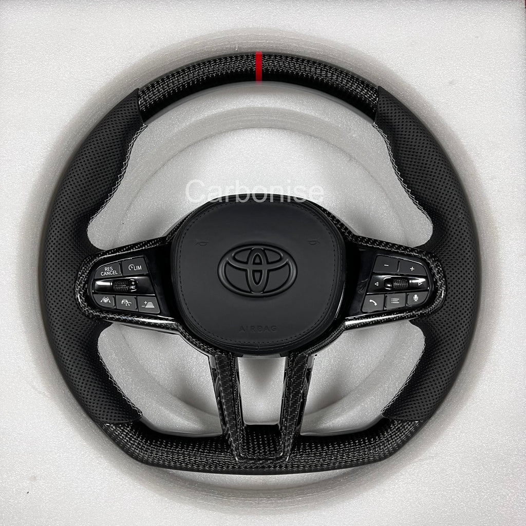 Carbonise Supra steering wheel with perforated leather grips, red center stripe, carbon fiber top and bottom, flat bottom design, and OEM multifunction buttons