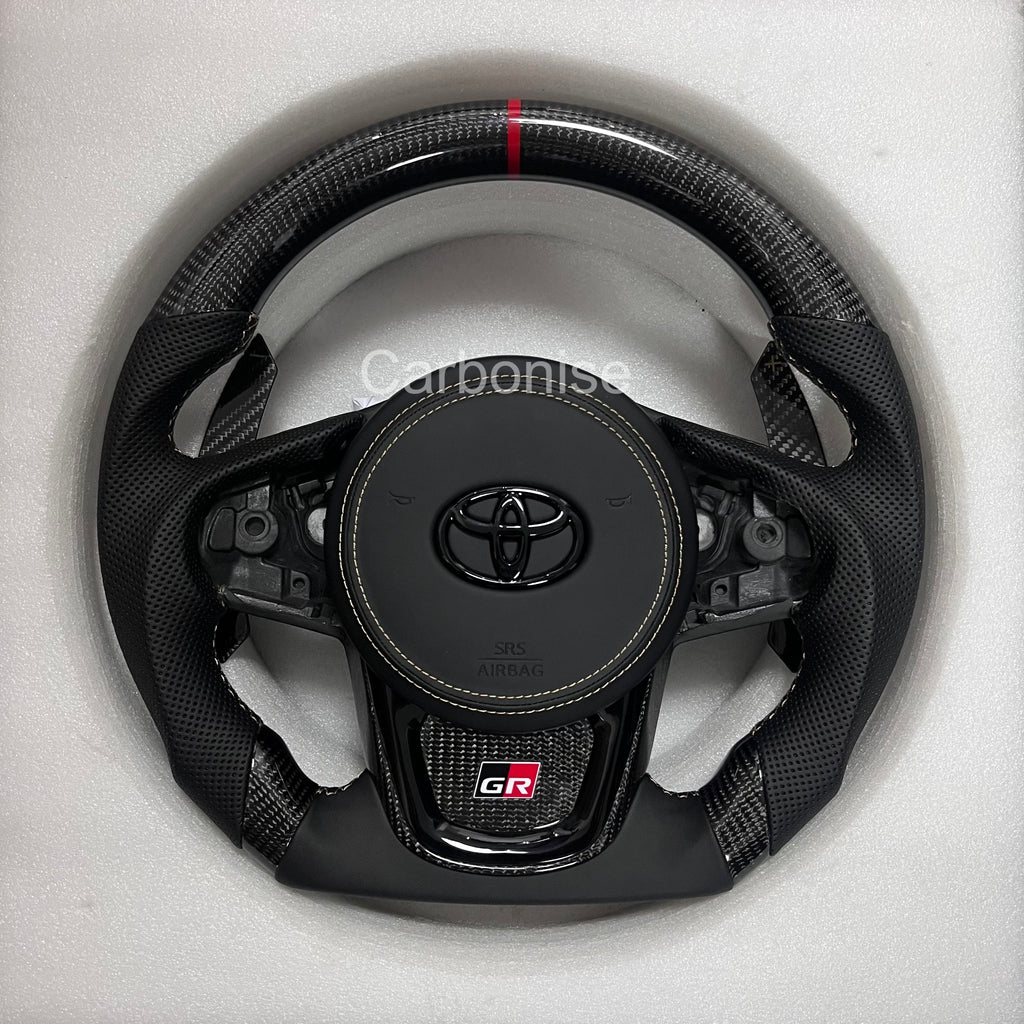 Carbonise Supra carbon fiber steering wheel with perforated leather grips, red center stripe, flat bottom design, GR badge, and OEM button layout