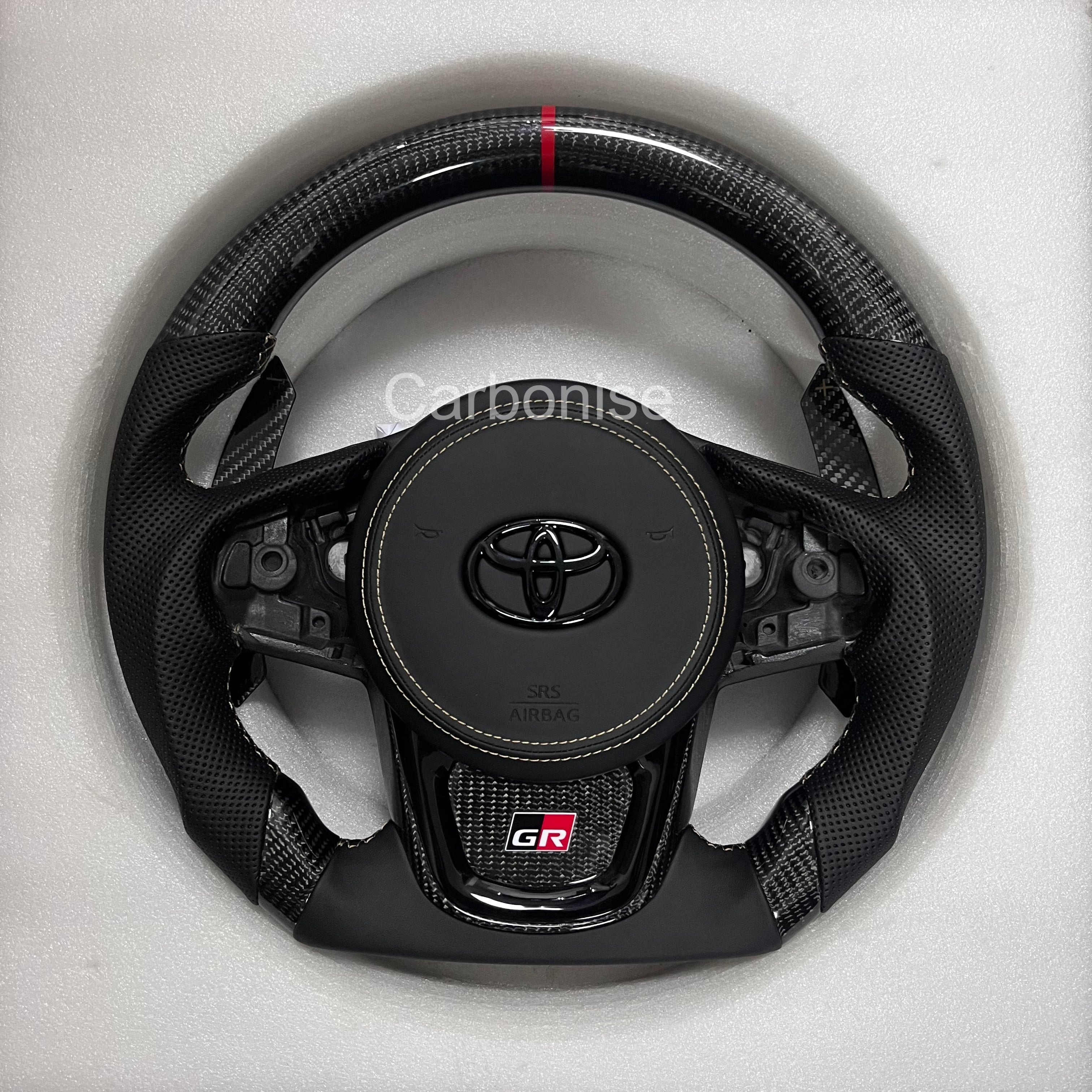 Carbonise Supra carbon fiber steering wheel with perforated leather grips, red center stripe, flat bottom design, GR badge, and OEM button layout