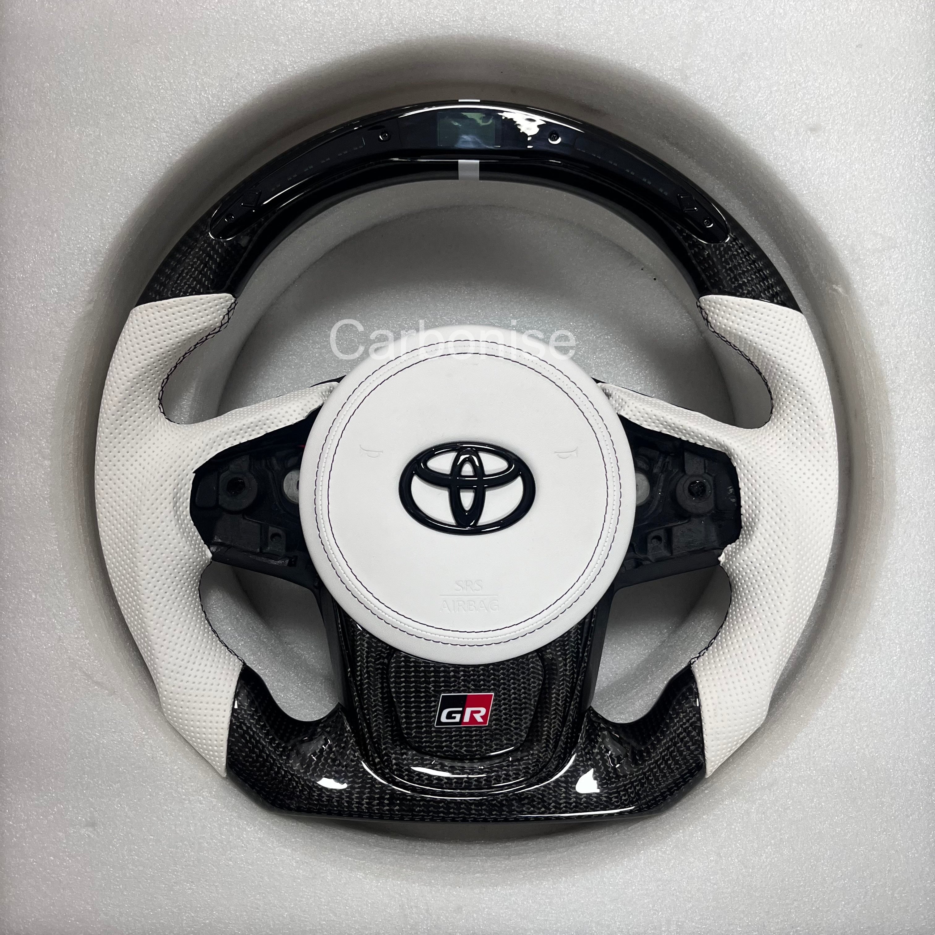 Carbonise Supra carbon fiber steering wheel with white perforated leather grips, white airbag cover, flat bottom design, GR badge, and OEM button layout, led shift light