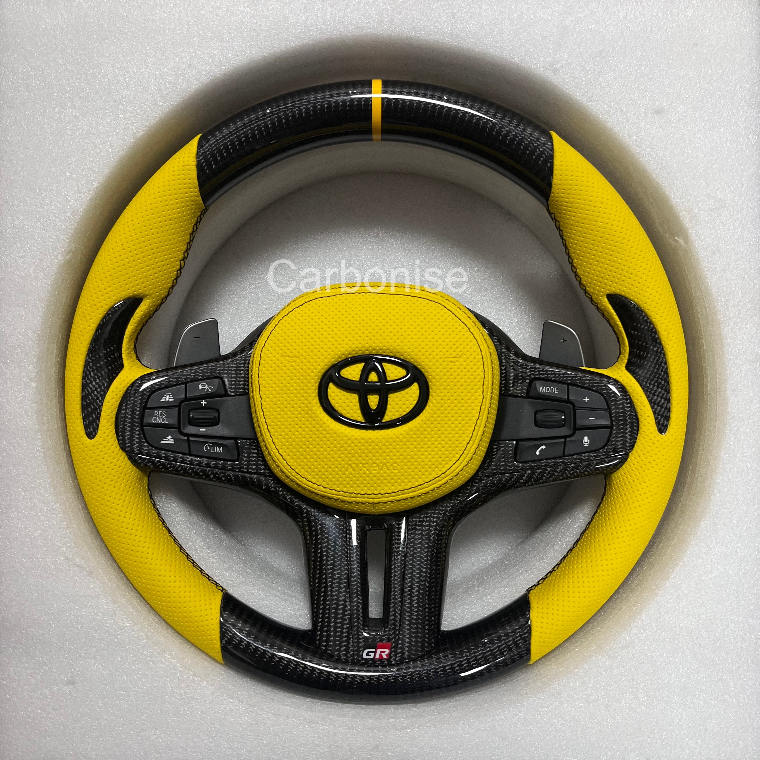 Custom Supra carbon fiber steering wheel with yellow perforated leather grips, yellow center stripe, carbon fiber top and bottom, and GR badge by Carbonise
