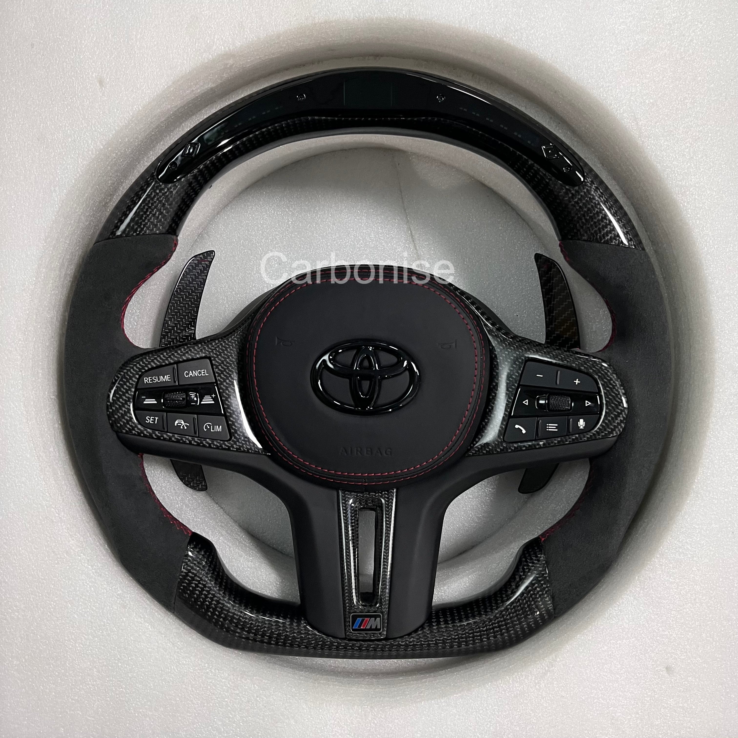 Custom Supra carbon fiber steering wheel with Alcantara grips, red stitching, carbon fiber top and bottom, multifunction buttons, and M-style badge by Carbonise