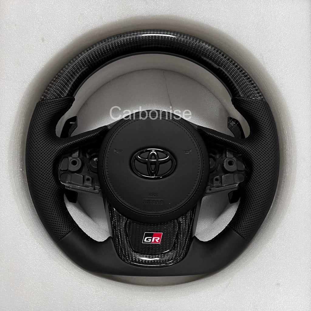 Supra carbon fiber steering wheel with gloss carbon top and bottom, perforated leather grips, flat bottom design, and OEM airbag compatibility.