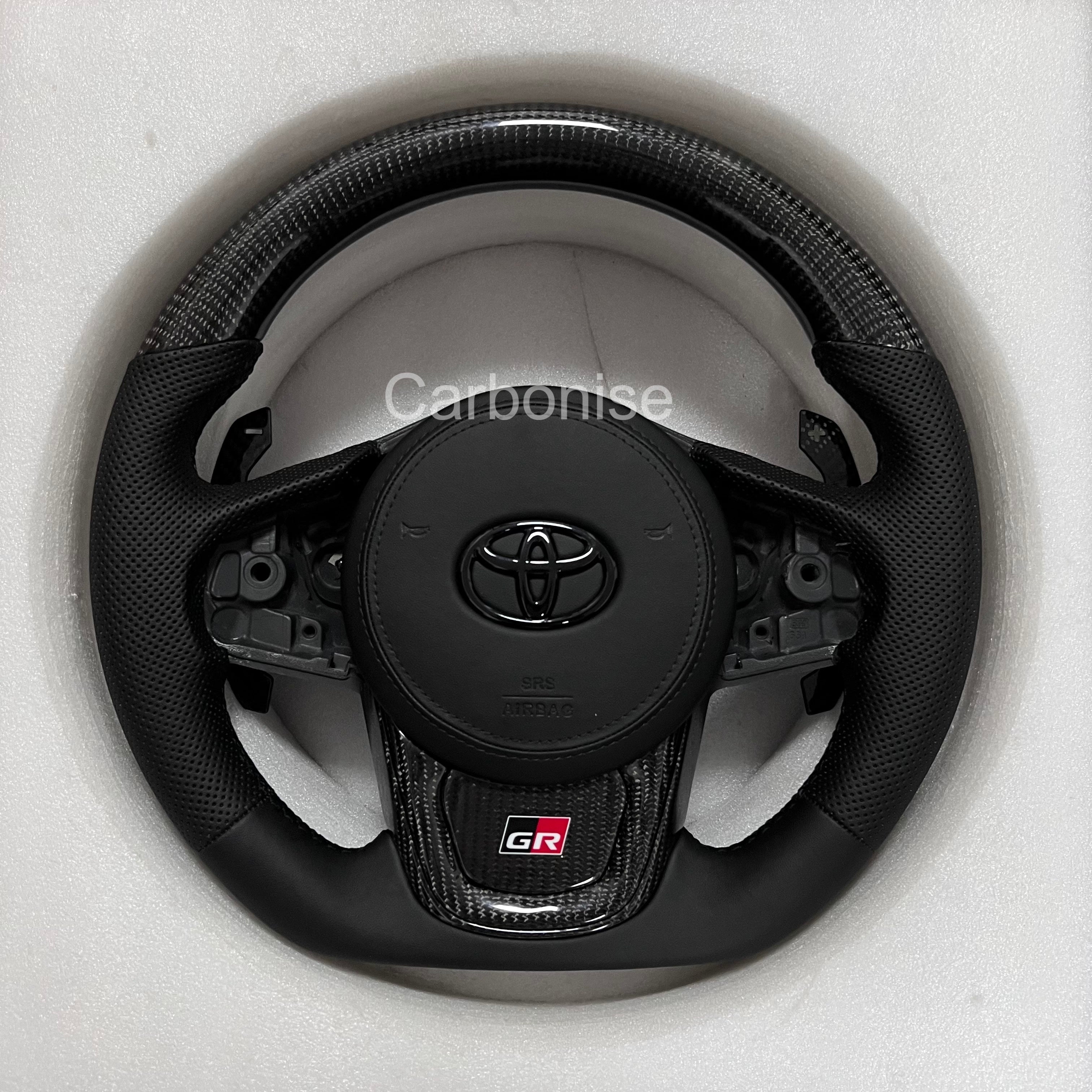 Supra carbon fiber steering wheel with gloss carbon top and bottom, perforated leather grips, flat bottom design, and OEM airbag compatibility.