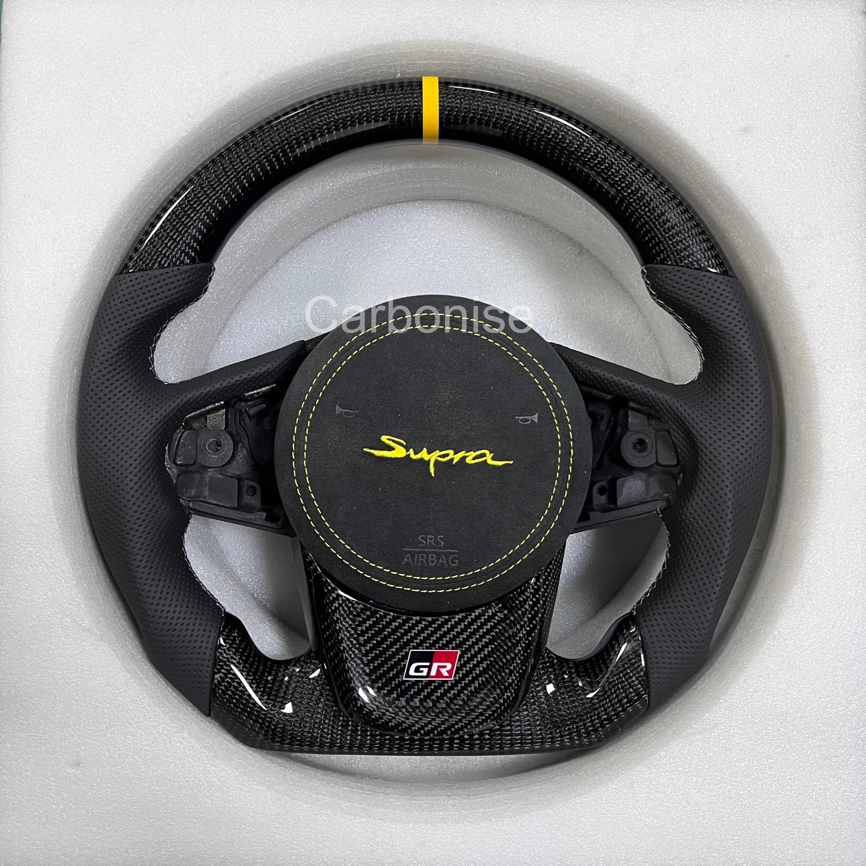 Toyota GR Supra carbon fiber steering wheel with yellow center stripe, perforated leather grips, flat bottom design, GR badge, and OEM airbag compatibility.