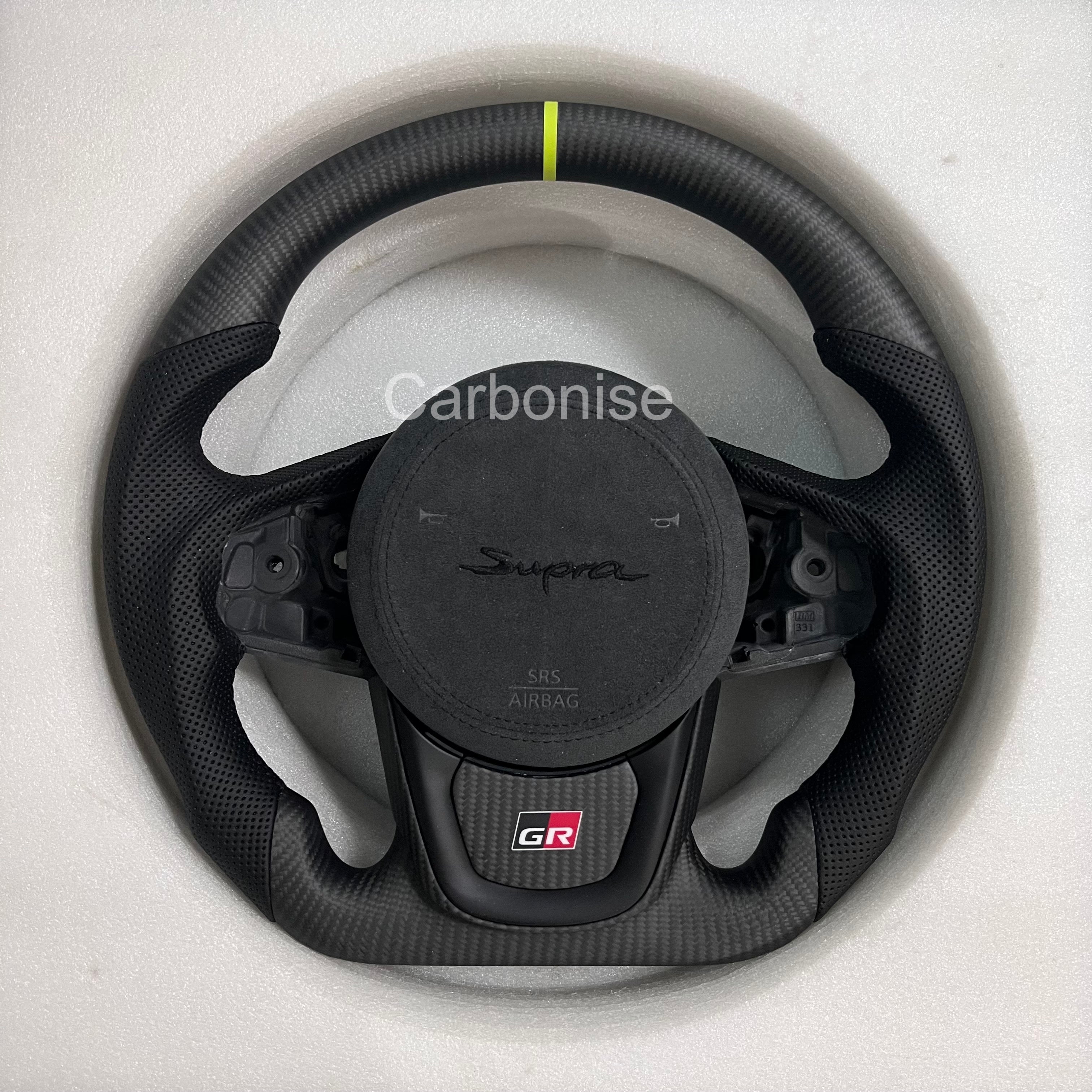 Toyota GR Supra carbon fiber steering wheel with neon green center stripe, perforated leather grips, flat bottom design, GR badge, and OEM airbag fitment.