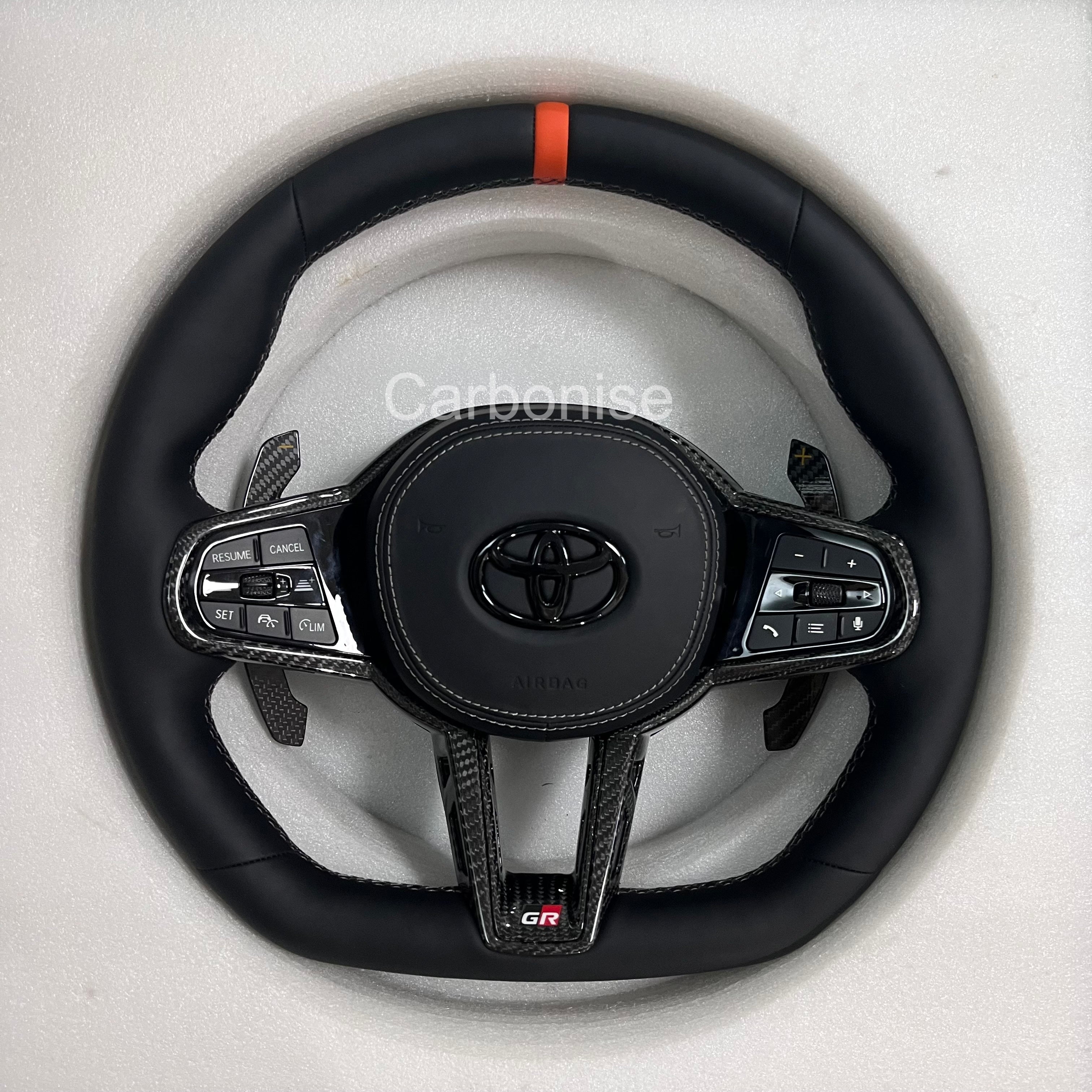 Supra steering wheel with orange center stripe, smooth leather grip, flat bottom design, carbon fiber trim, paddle shifters, and OEM airbag fitment.
