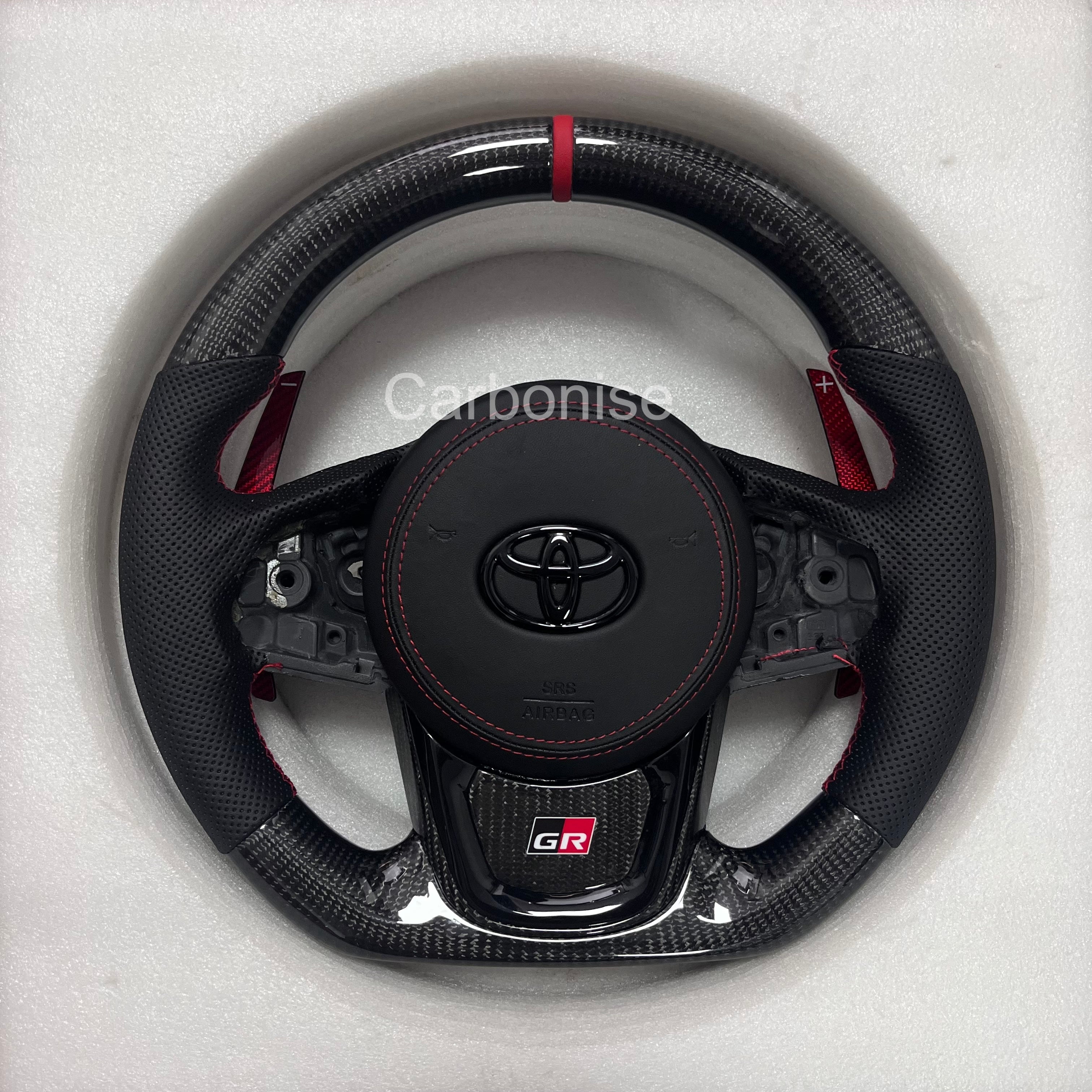 Supra carbon fiber steering wheel with red center stripe, perforated leather grip, flat bottom design, paddle shifters, and OEM airbag fitment.
