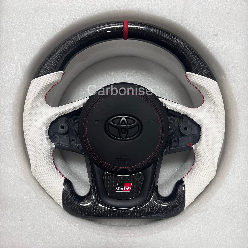 Custom Supra steering wheel featuring a carbon fiber top and bottom, white perforated leather side grips, red center stripe, red stitching, flat bottom design, and GR badge.