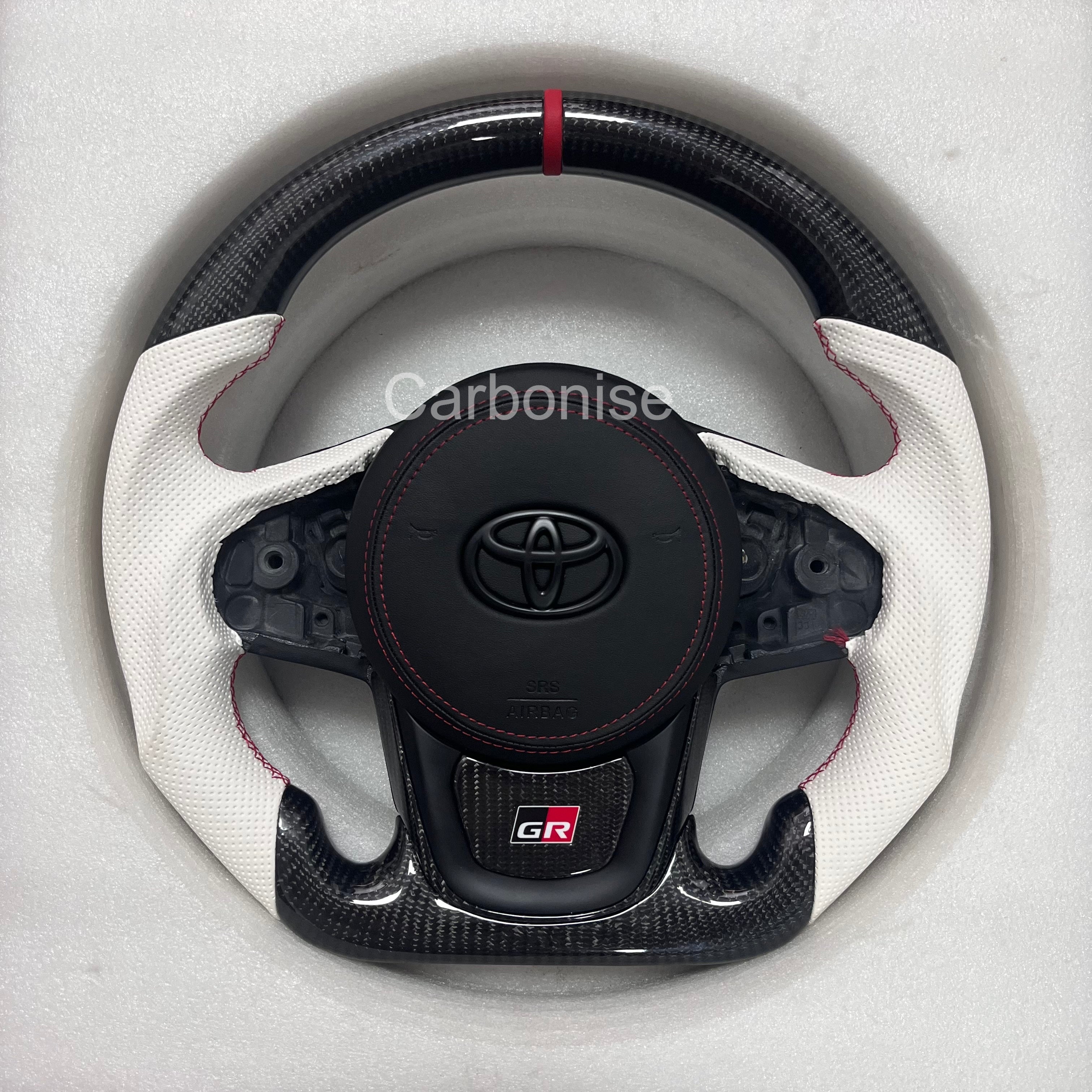 Custom Supra steering wheel featuring a carbon fiber top and bottom, white perforated leather side grips, red center stripe, red stitching, flat bottom design, and GR badge.