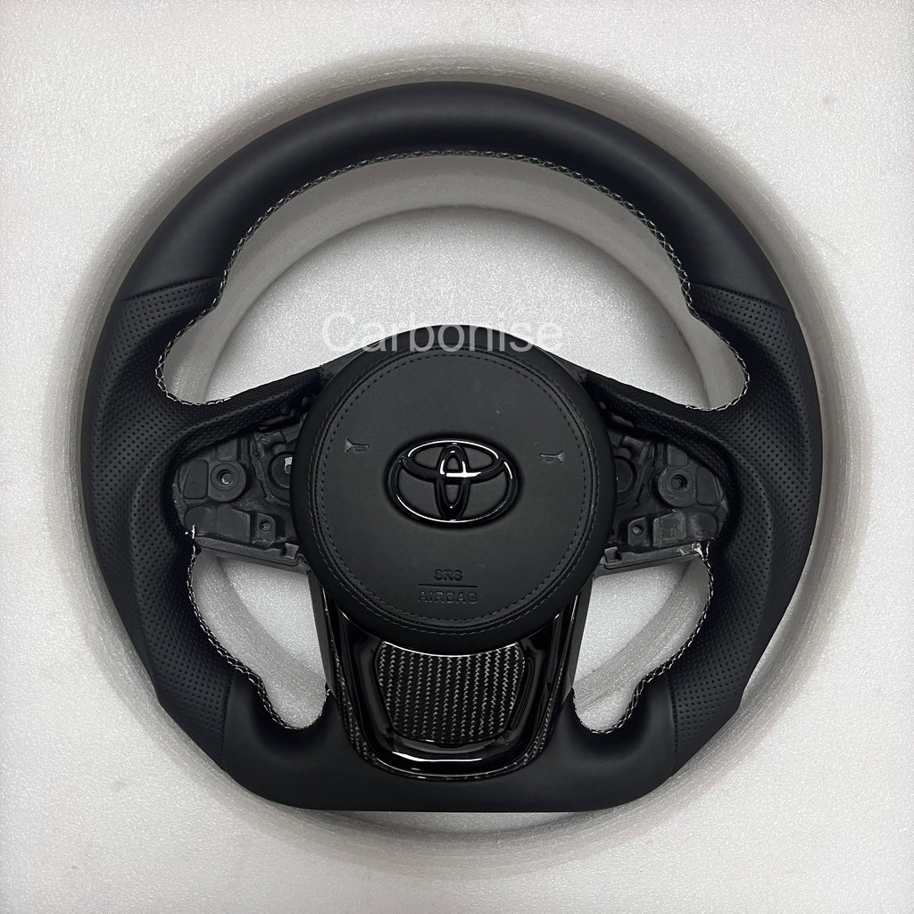 Supra steering wheel featuring full black leather wrap with perforated side grips, smooth leather top section, OEM multifunction controls, and factory-style center airbag.