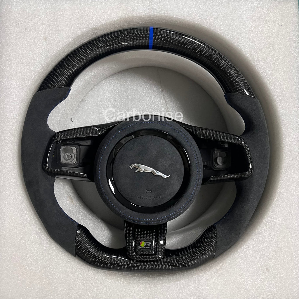 Custom Jaguar R carbon fibre steering wheel with Alcantara side grips, gloss carbon trim, blue centre alignment stripe, and flat-bottom sport design.