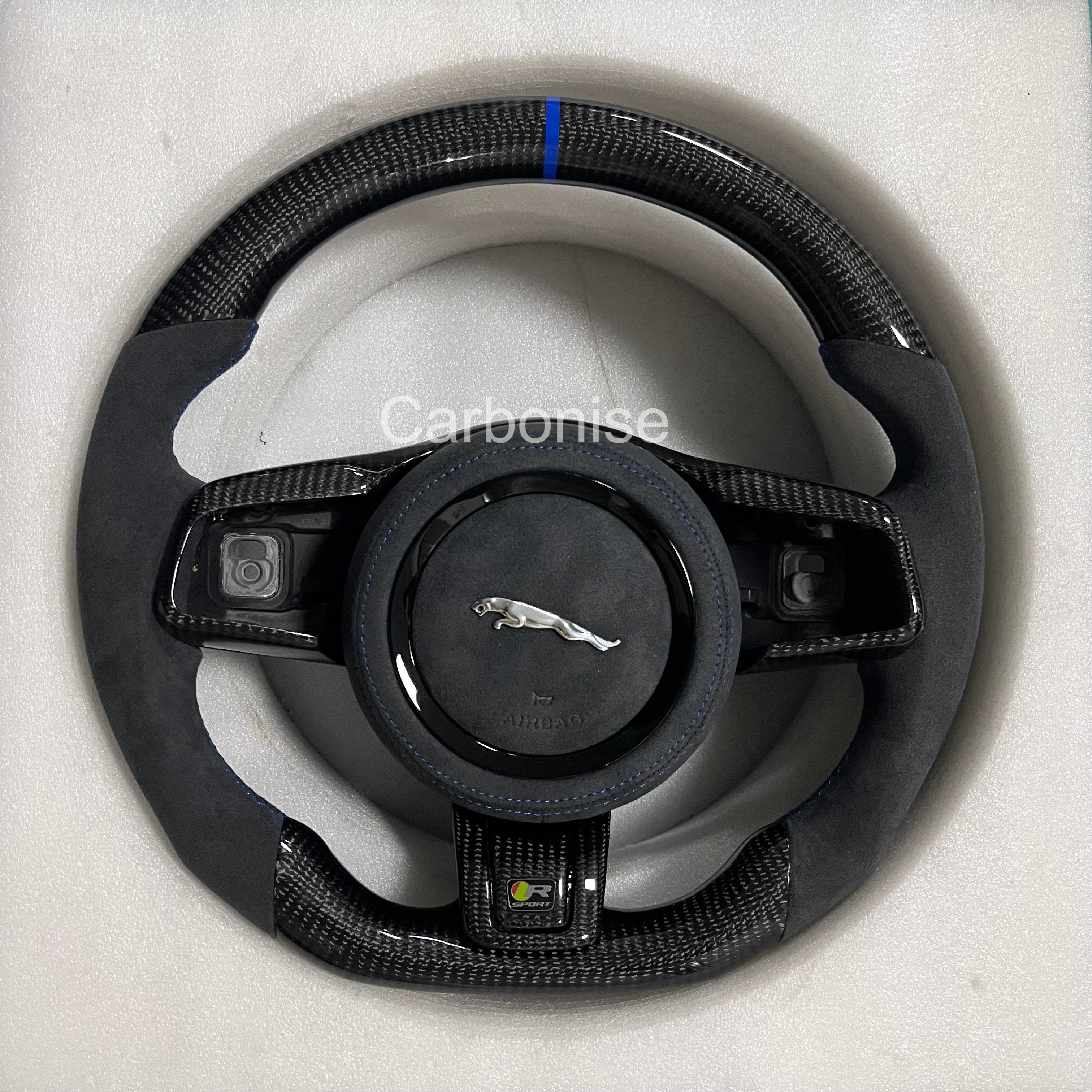 Custom Jaguar R carbon fibre steering wheel with Alcantara side grips, gloss carbon trim, blue centre alignment stripe, and flat-bottom sport design.