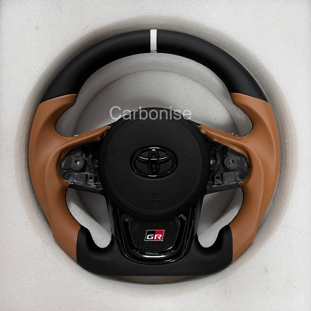 Custom Supra steering wheel with brown leather side grips, black leather top and bottom, white top marker, and carbon fibre lower spoke.