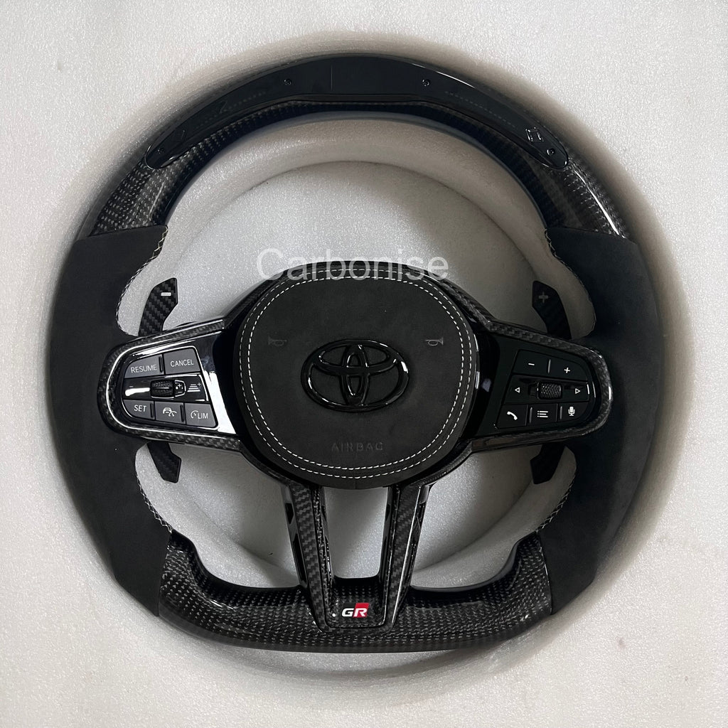 Custom Supra steering wheel featuring carbon fiber top and bottom, black Alcantara side grips, white contrast stitching, integrated paddle shifters, flat bottom design, and GR badge.