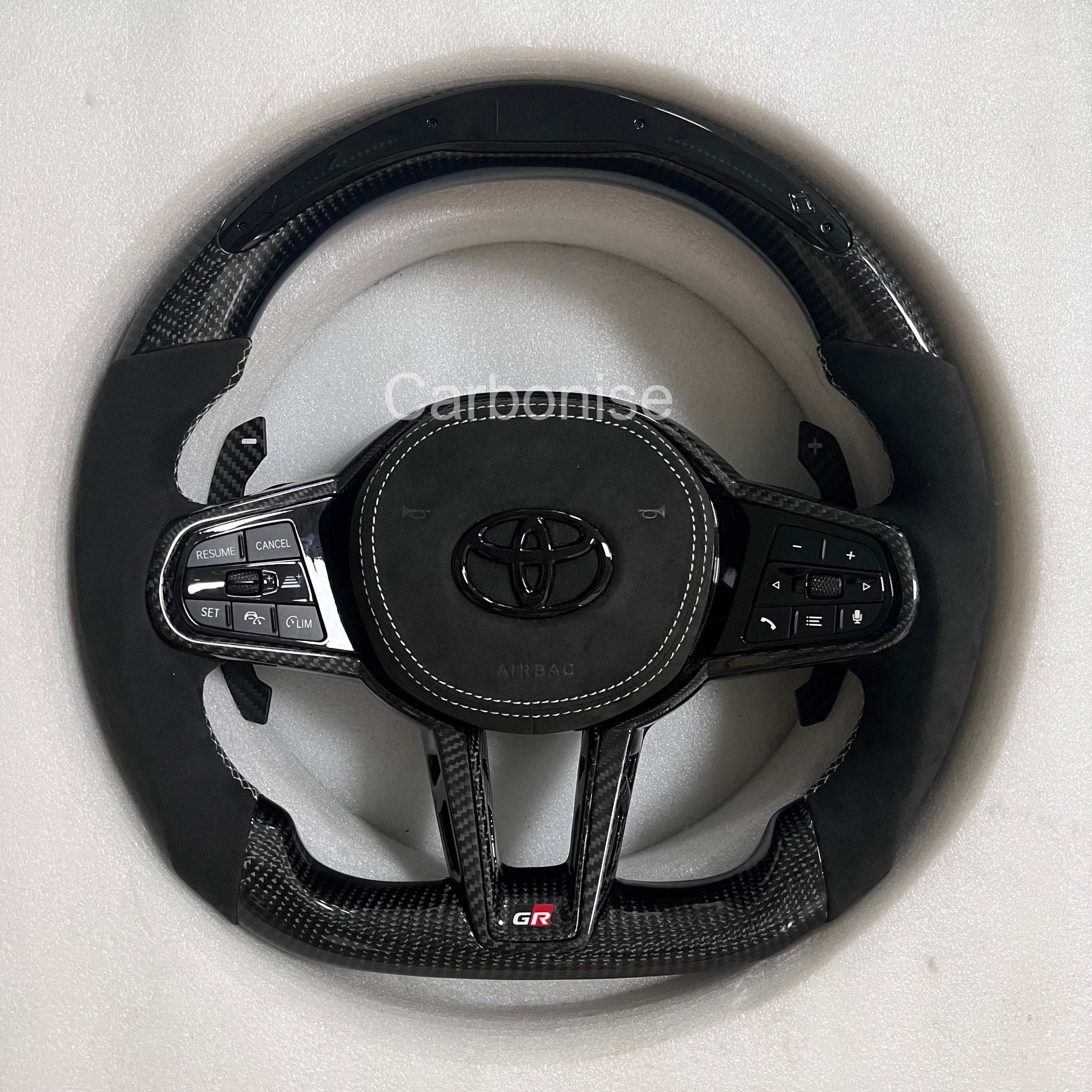 Custom Supra steering wheel featuring carbon fiber top and bottom, black Alcantara side grips, white contrast stitching, integrated paddle shifters, flat bottom design, and GR badge.